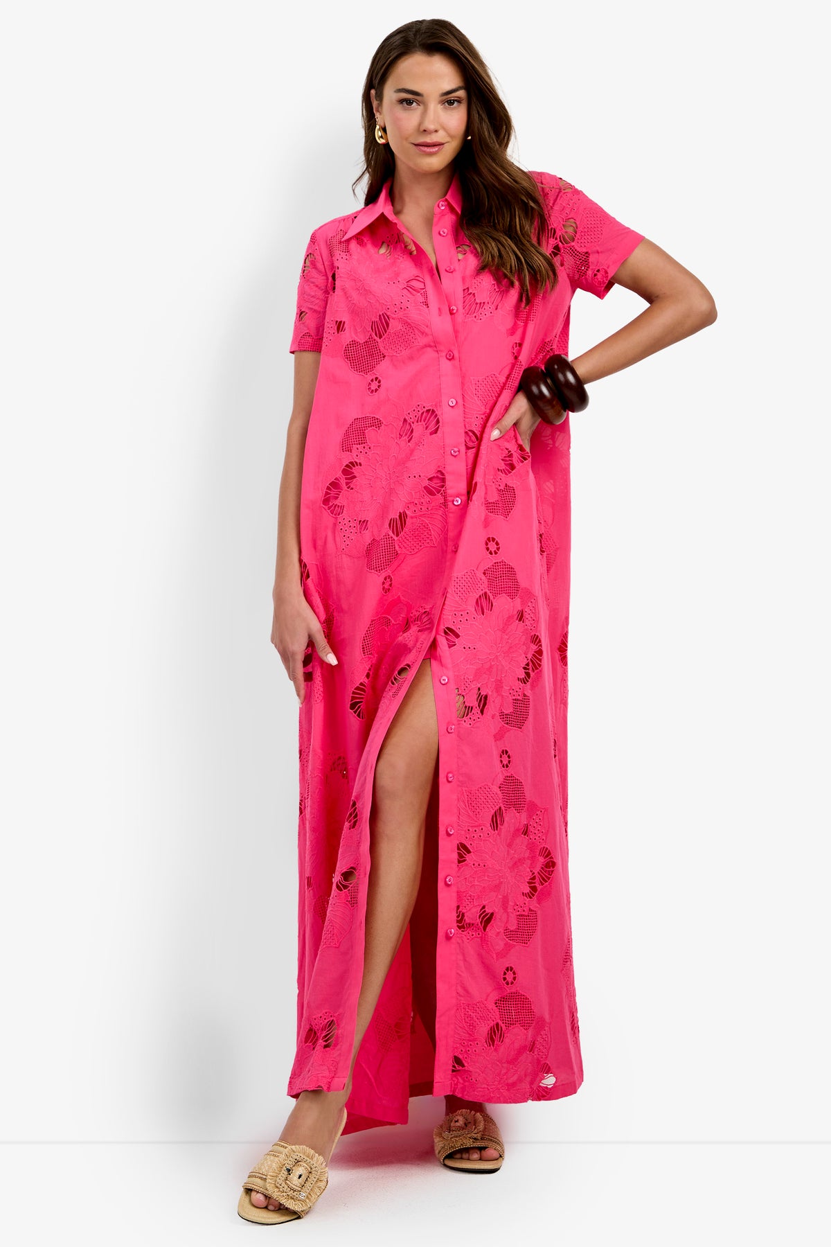 A woman wears the Kon Tiki Maxi Dress—a bright pink, short-sleeve, button-down maxi dress with a floral pattern—as she stands with one hand on her hip, accessorized with large bangles and beige sandals.