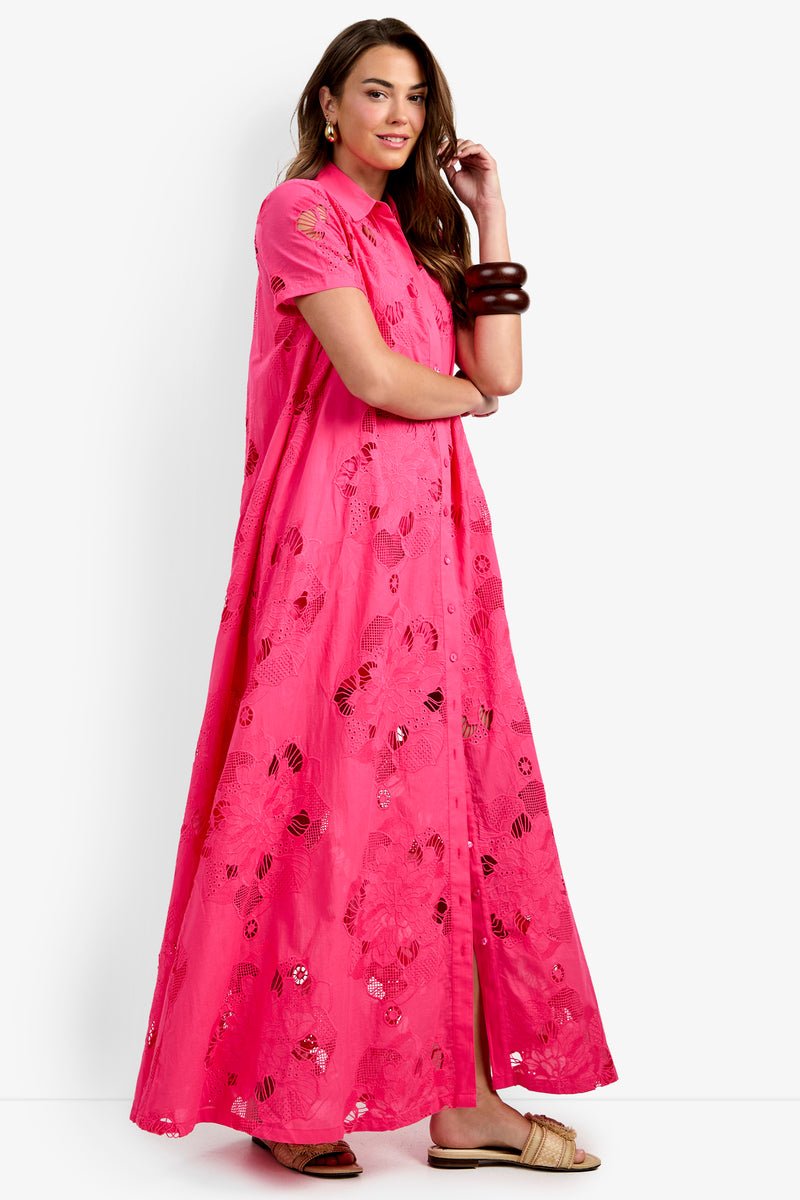 A woman wearing the Kon Tiki Maxi Dress—a bright pink, ankle-length dress with short sleeves and floral embroidery—paired with two chunky brown bracelets and tan sandals, stands against a white background.