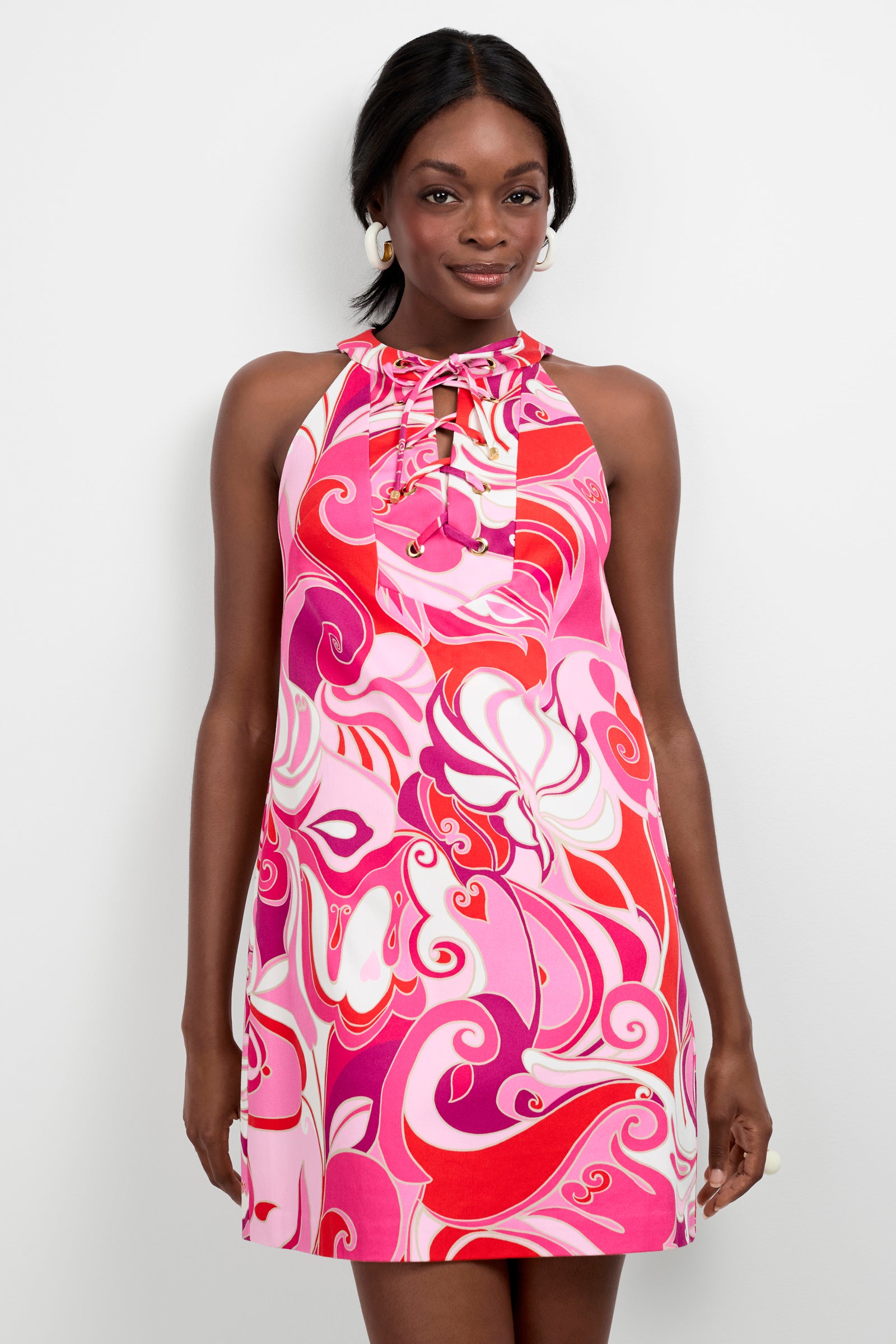 A woman models the Cappuccia Lace-Up Mini Dress, a sleeveless halter-neck style featuring a bold swirling pink, red, and white pattern with lace-up neckline detail. She poses against a plain white background, smiling softly.