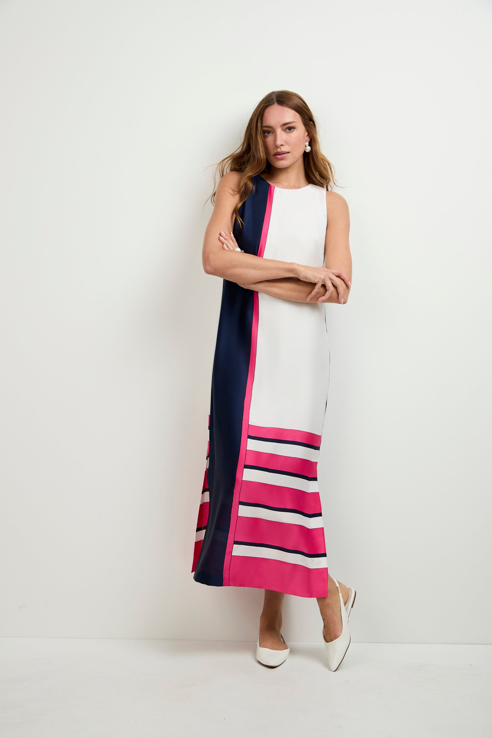 A woman with long hair stands against a white wall, wearing the Moderna Linee Midi Dress—a sleeveless dress with navy, white, and pink color blocks and stripes—paired with white pointed-toe shoes.