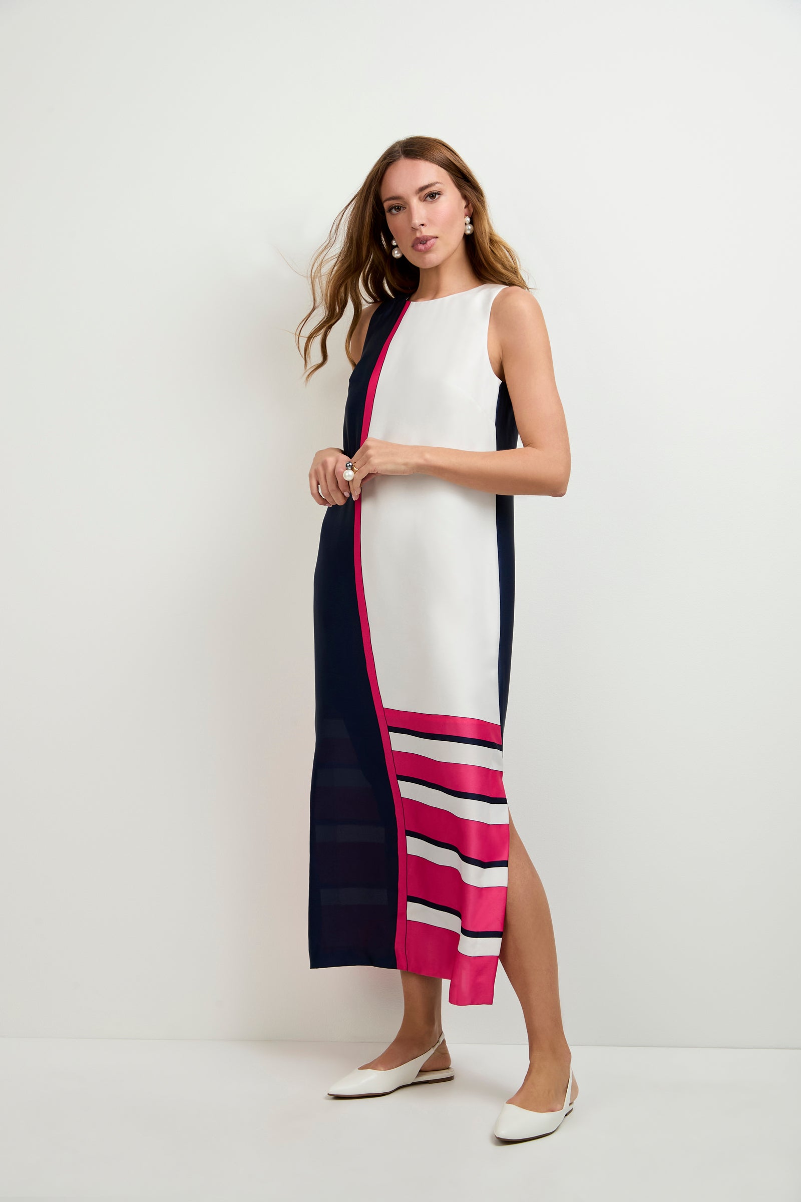 A woman stands against a plain white wall wearing the Moderna Linee Midi Dress, a sleeveless, long color-block dress in navy, white, and hot pink with pink and white stripes near the hem. She pairs it with white pointed flats.