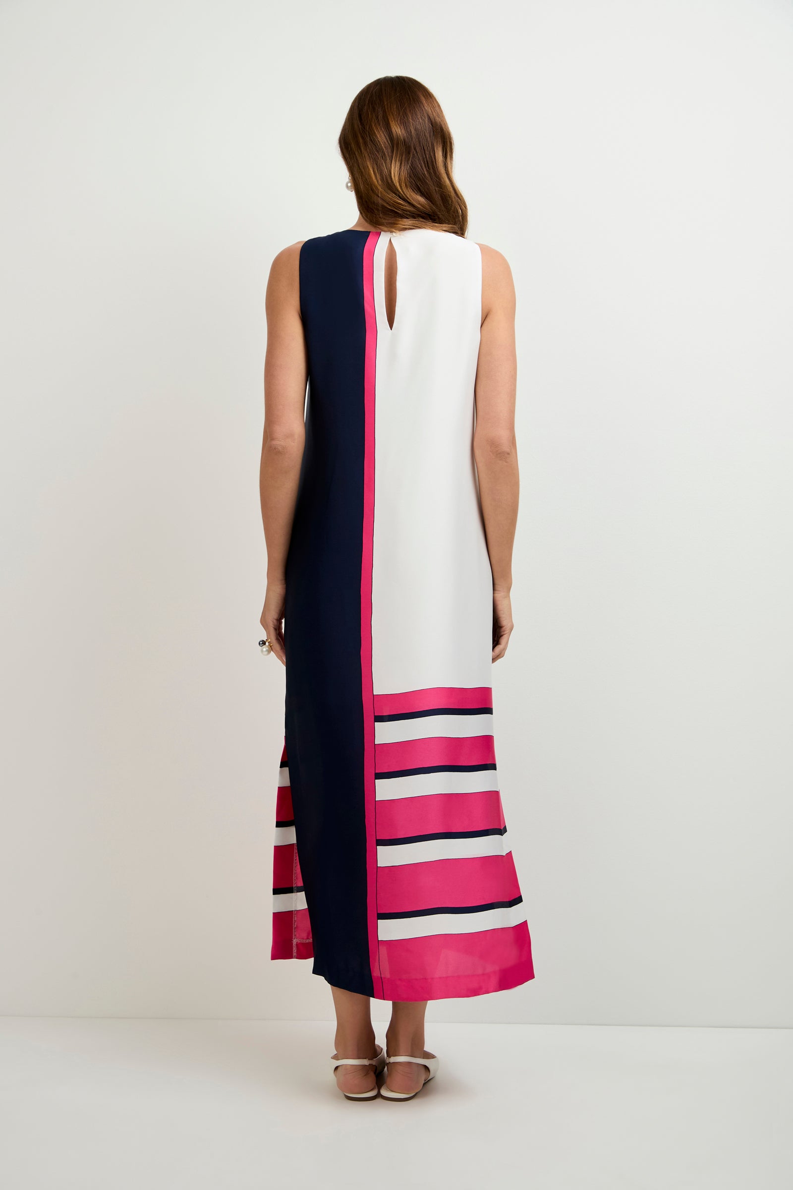 A woman with long brown hair wears the Moderna Linee Midi Dress—a sleeveless design with one navy side, one white side, and bold pink stripes near the hem. She pairs it with flat sandals against a plain white background.