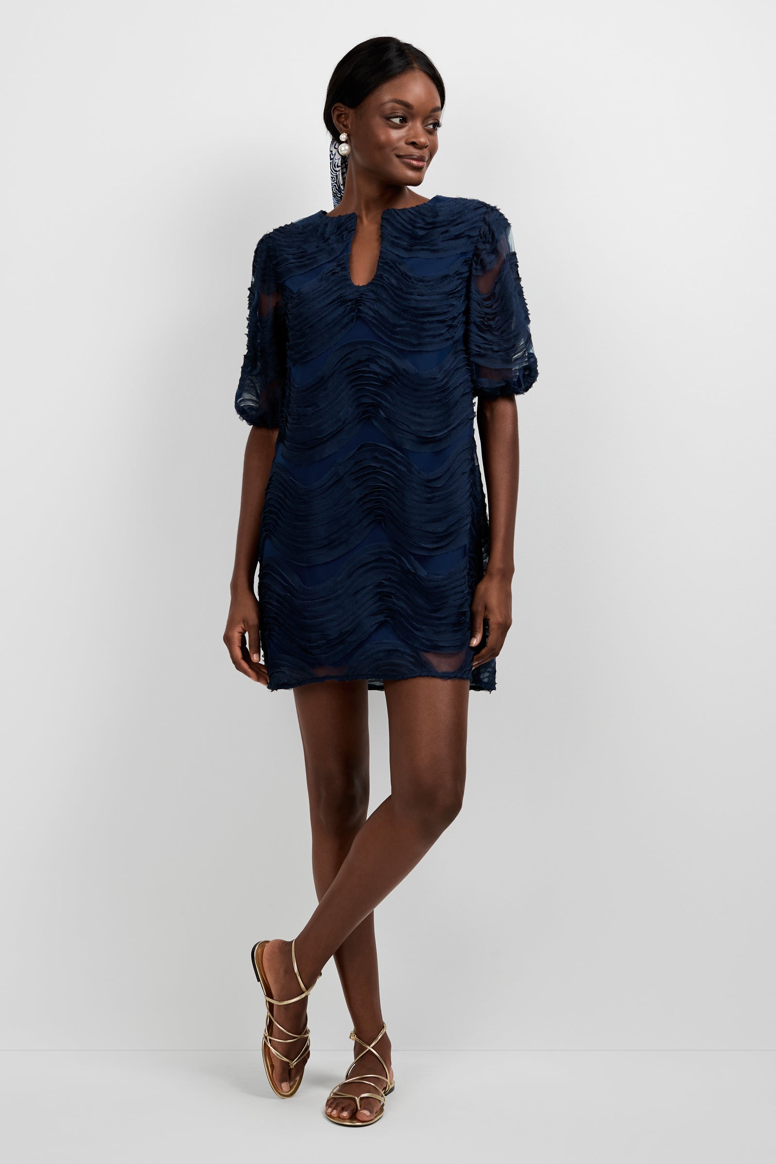 A woman poses against a plain background wearing the Torchino Organza Mini Dress, a short textured navy blue dress with short sleeves and a keyhole neckline, styled with gold strappy flat sandals and statement earrings.