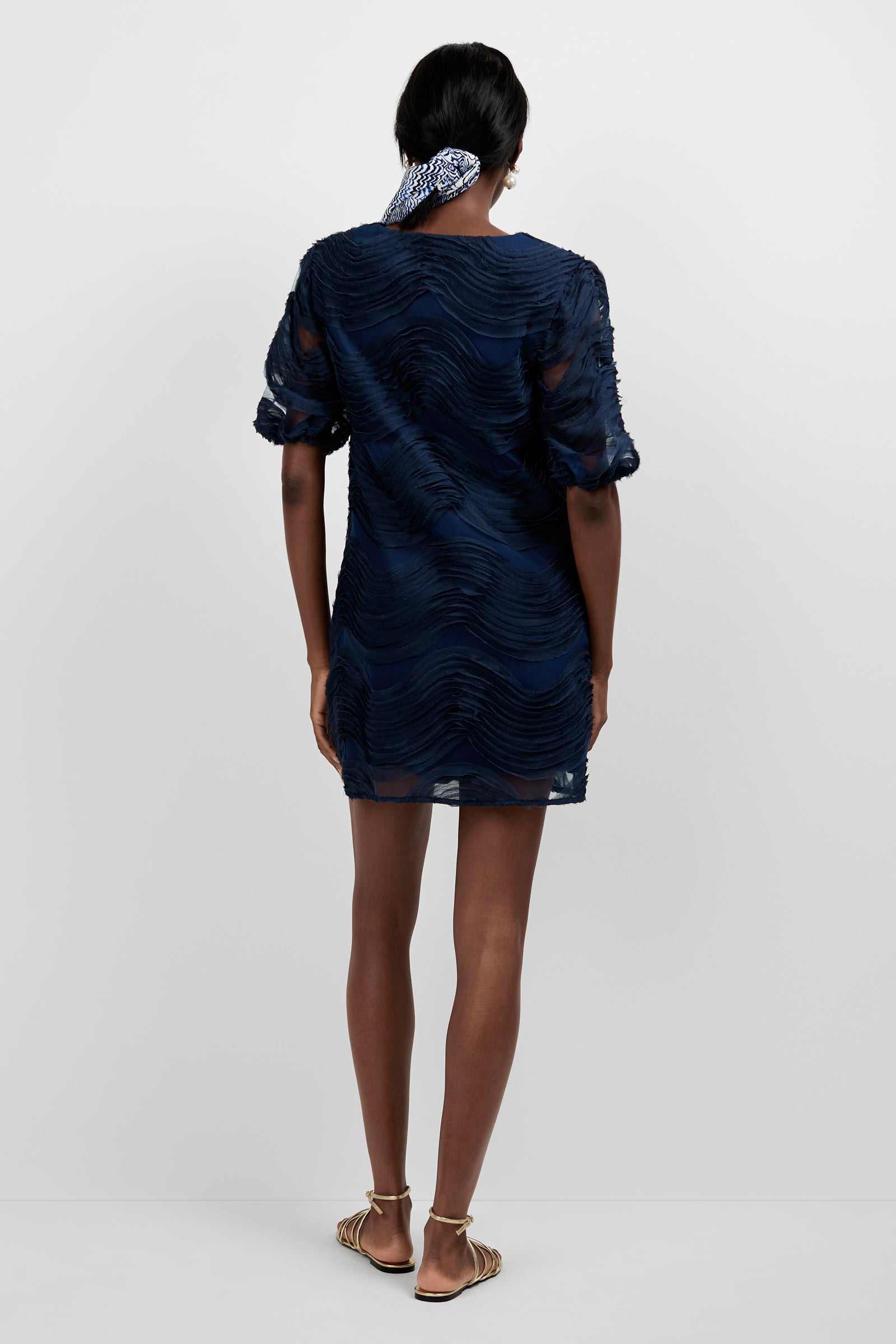 A woman faces away in the Torchino Organza Mini Dress—short, textured, and navy blue with puff sleeves. She wears flat sandals and a black-and-white patterned hair scarf against a plain white background.