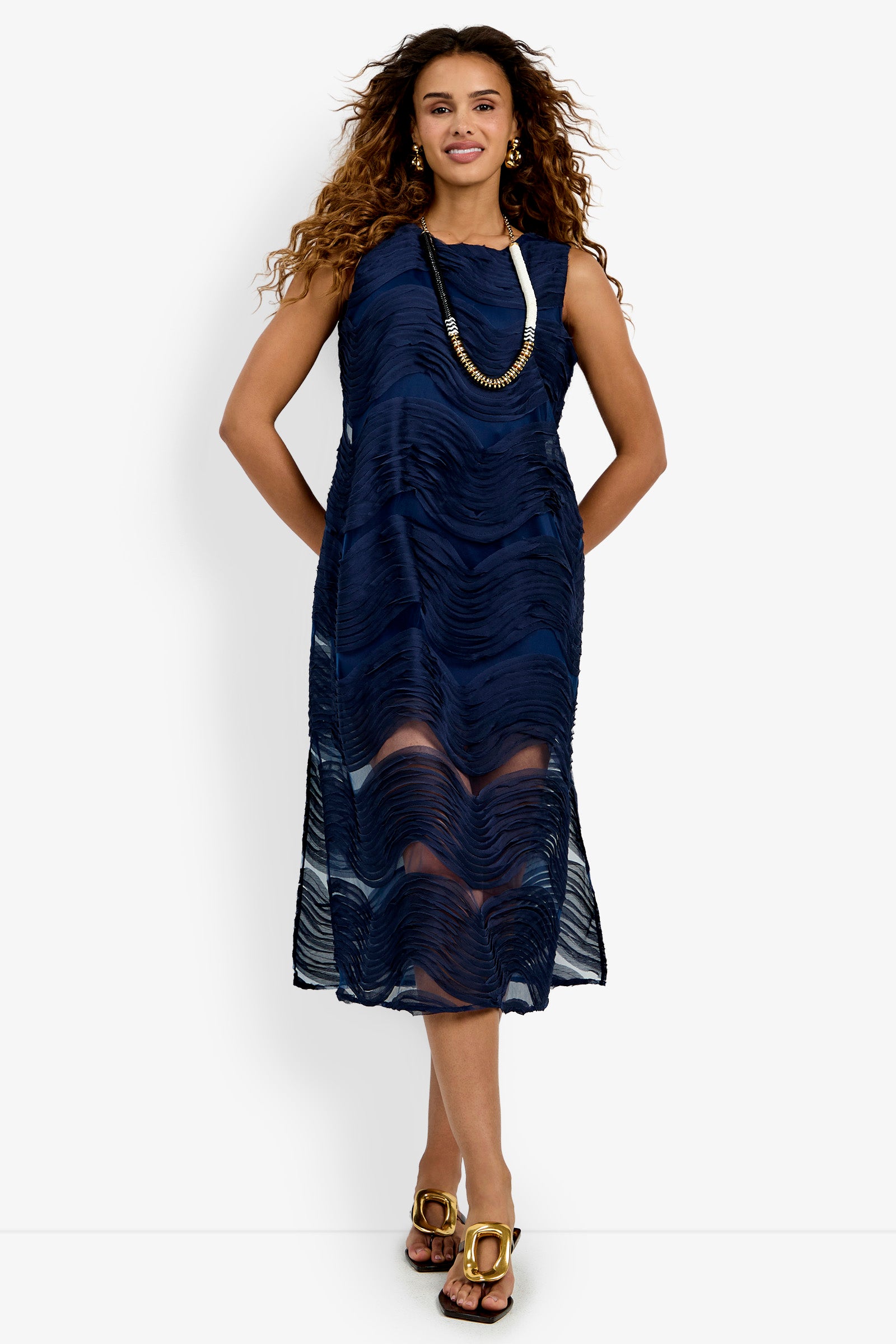 Wearing the Belisama Organza Midi Dress—a sleeveless, navy blue, textured midi dress with zigzag patterns—a woman pairs it with gold sandals and a long black necklace, standing and smiling against a white background.