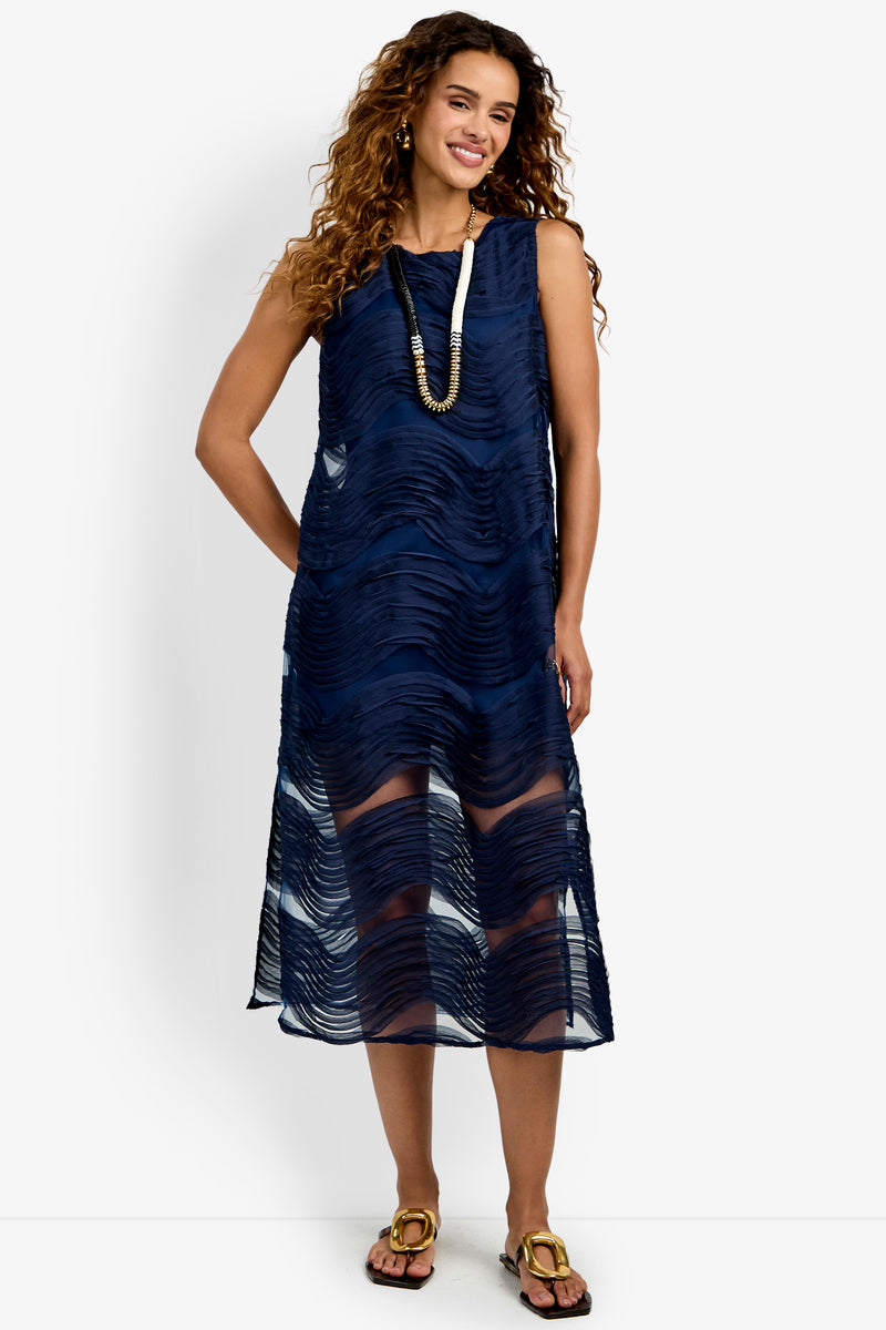 A woman stands and smiles against a white background, wearing the Belisama Organza Midi Dress—a sleeveless navy blue dress featuring textured wavy patterns and layered over a slip. She pairs the look with sandals, a long necklace, and has curly hair.
