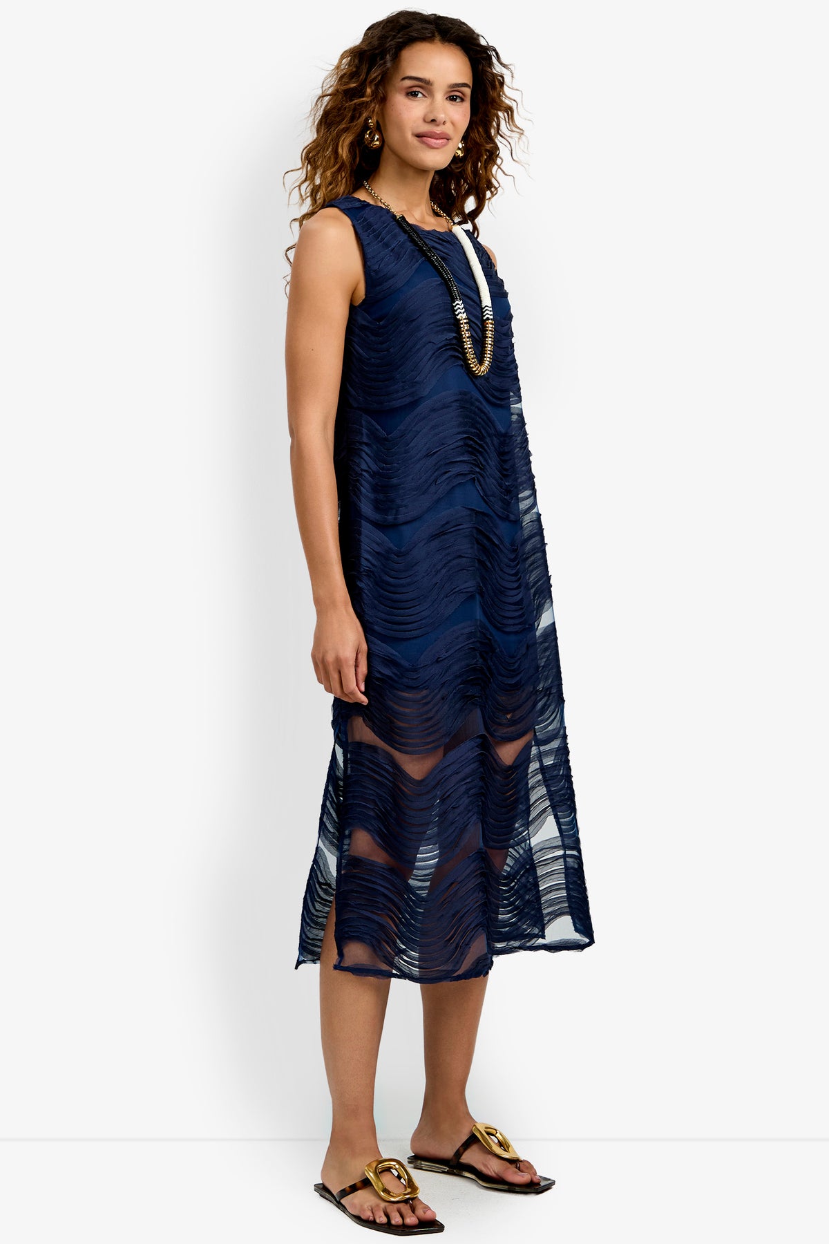 A woman wearing the Belisama Organza Midi Dress—a sleeveless, knee-length navy blue dress with a wavy textured pattern—paired with a long necklace and black sandals, standing and smiling.