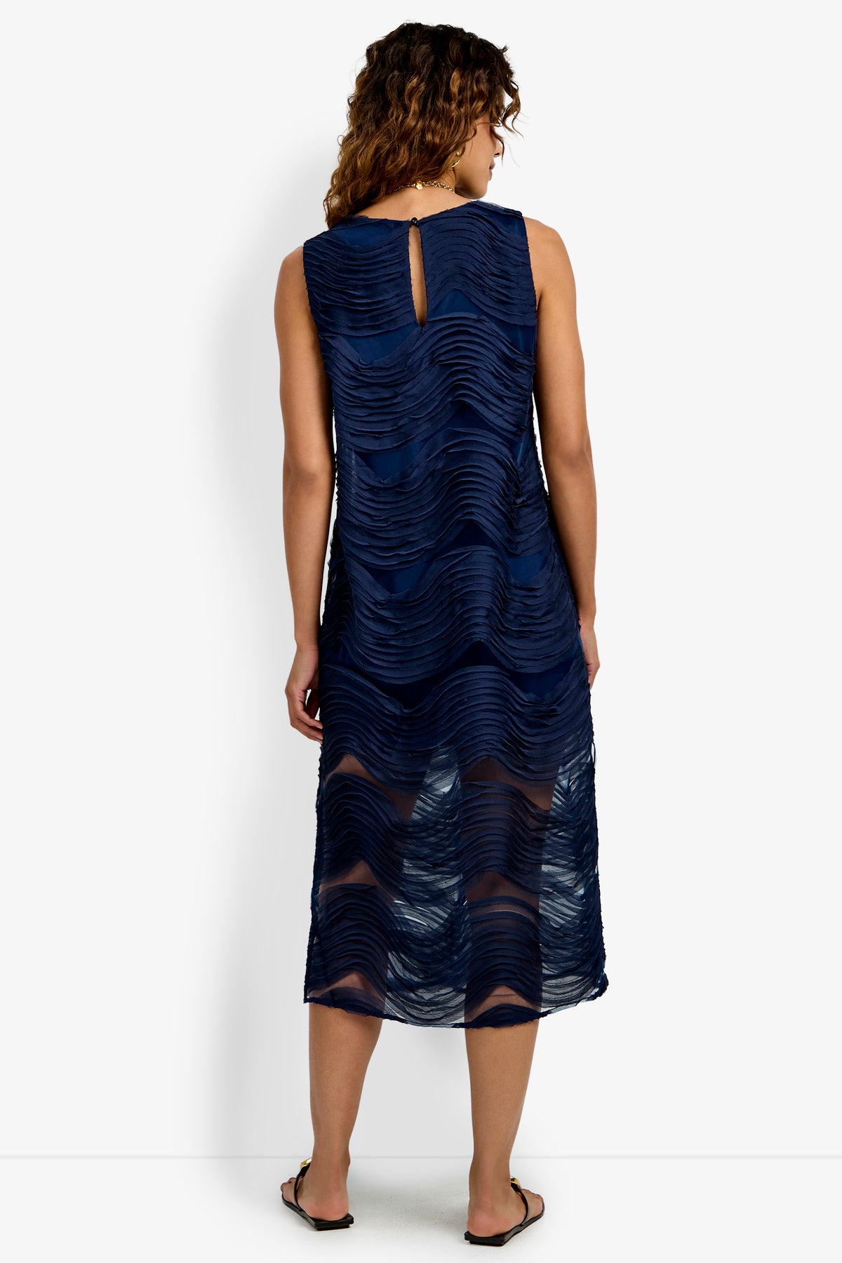 A person with curly hair wears the Belisama Organza Midi Dress—a sleeveless, navy blue, midi-length dress featuring a textured wavy pattern and a keyhole back—standing in sandals and facing away.