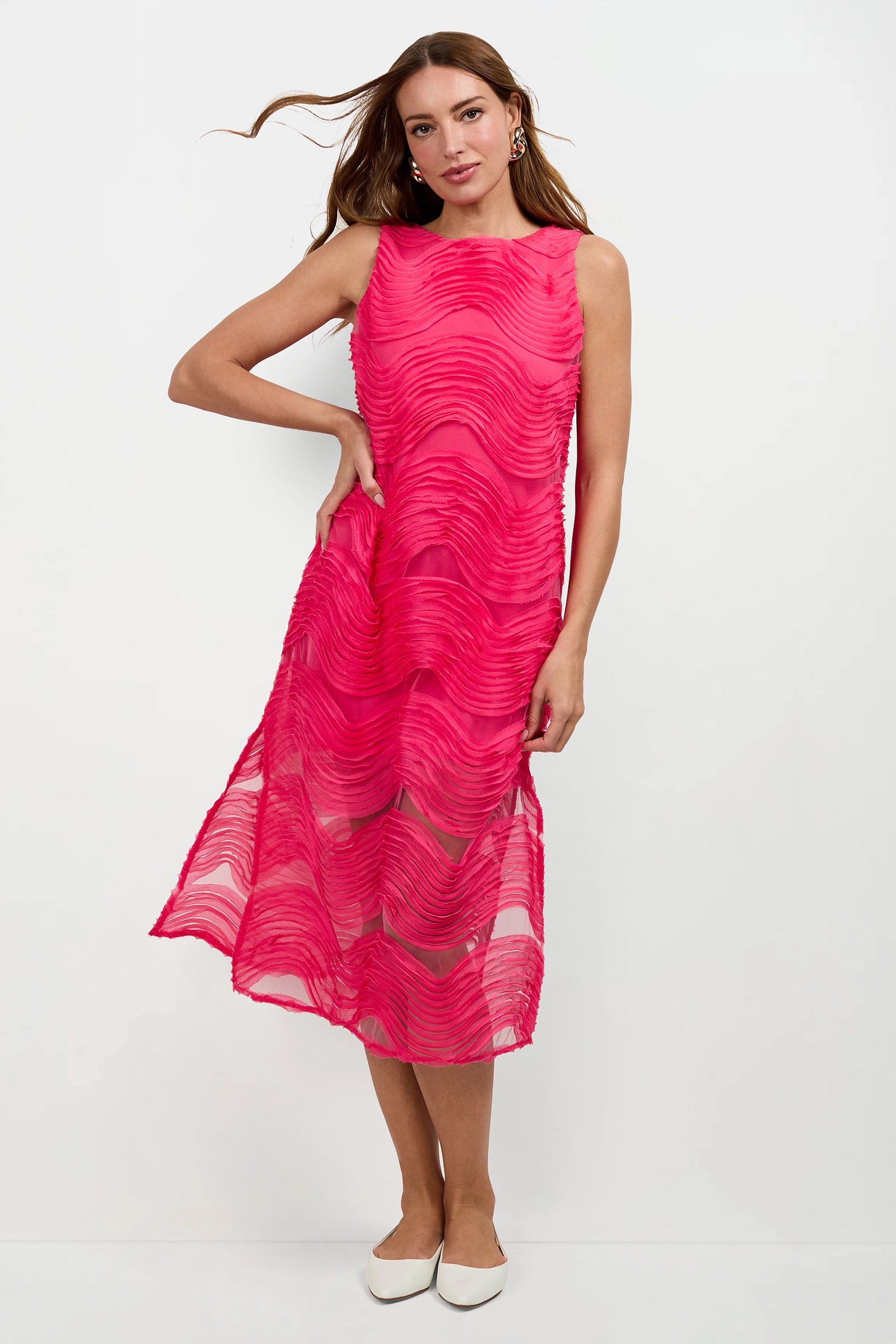 A woman stands against a plain background wearing the Belisama Organza Midi Dress—sleeveless, bright pink with a wavy texture and sheer overlay—paired with white pointed flats. Her hair is loose and one hand rests on her hip.