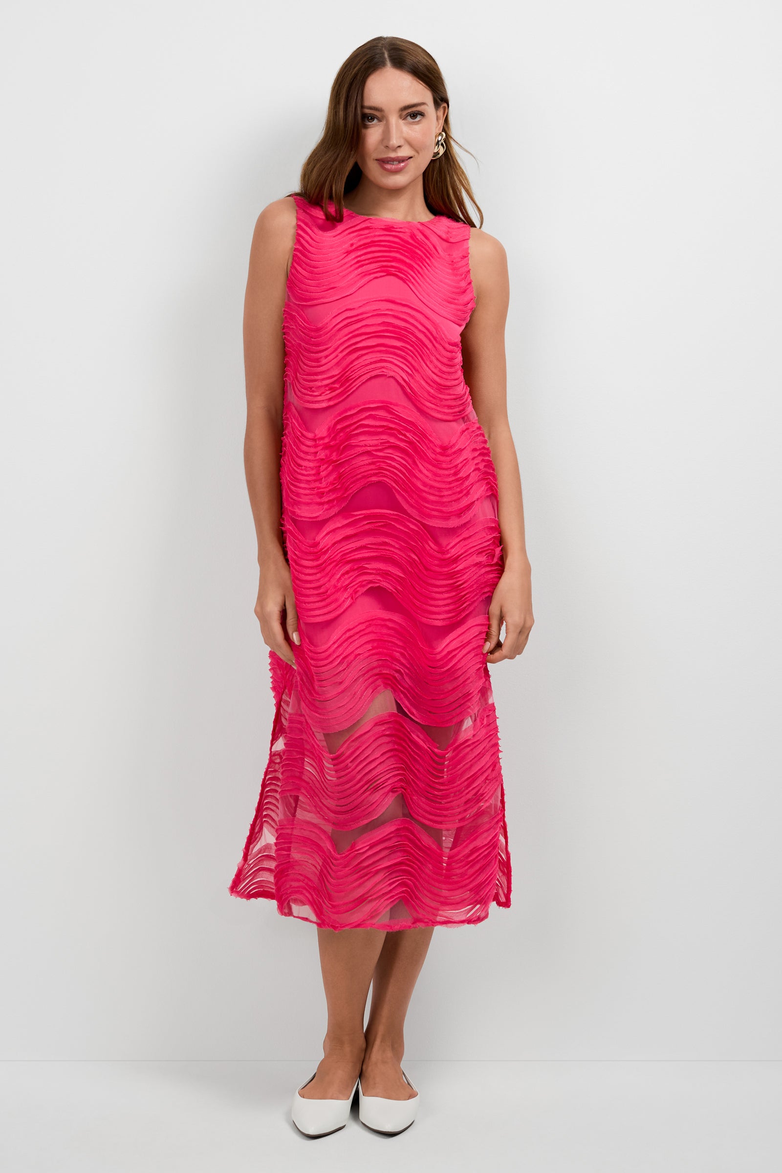 A woman with long hair and a gentle smile wears the Belisama Organza Midi Dress—sleeveless, ankle-length, pink with wavy textured patterns—paired with white pointed-toe shoes against a white background.
