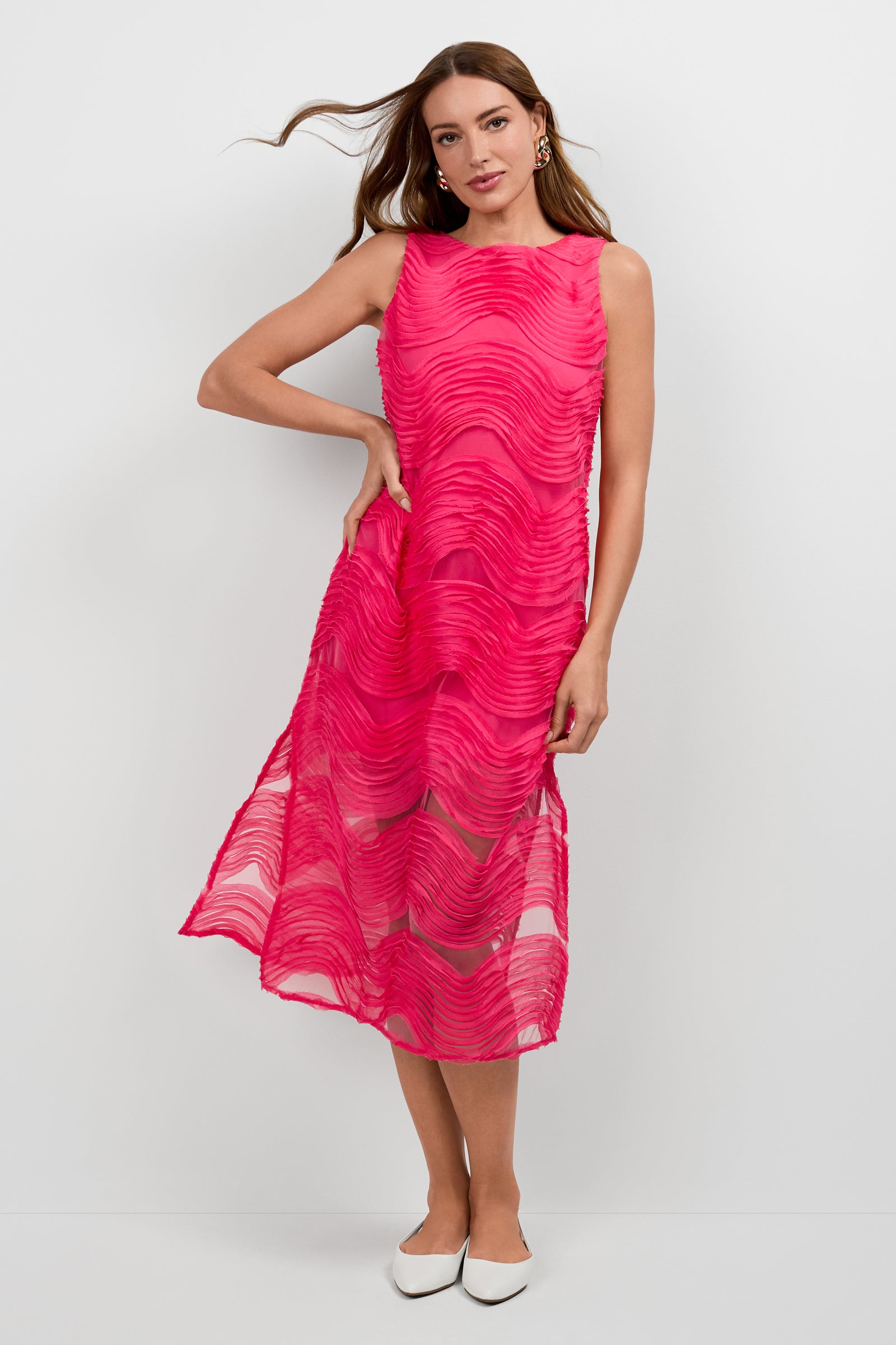 A woman stands against a plain background, wearing the sleeveless Belisama Organza Midi Dress in bright pink with wavy textured patterns and sheer sections. She styles it with loose hair, gold earrings, and white pointed-toe flats.