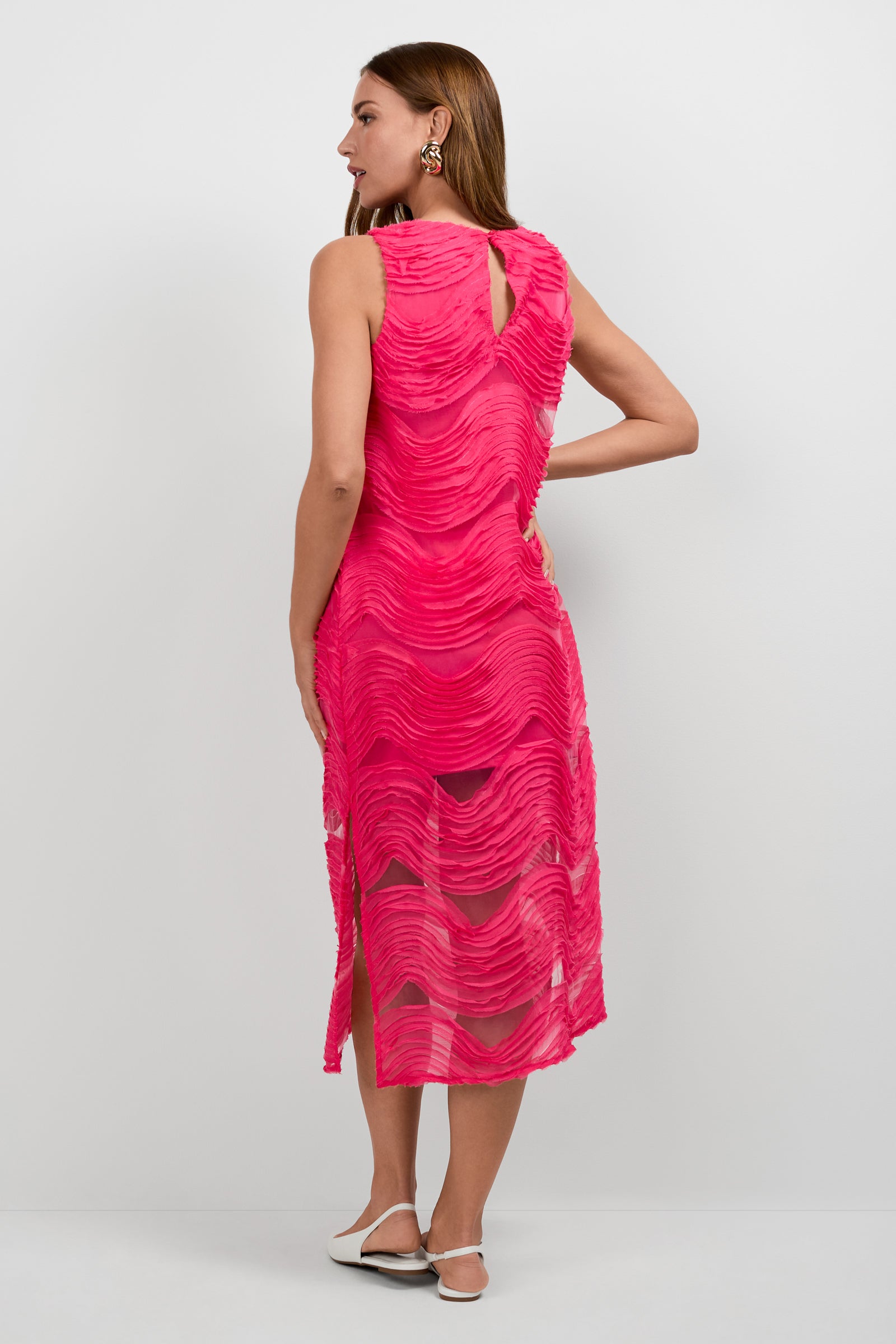 A woman with straight, light brown hair wears the Belisama Organza Midi Dress—a sleeveless, knee-length bright pink dress featuring wavy textured patterns and sheer panels—paired with white flats and statement earrings.