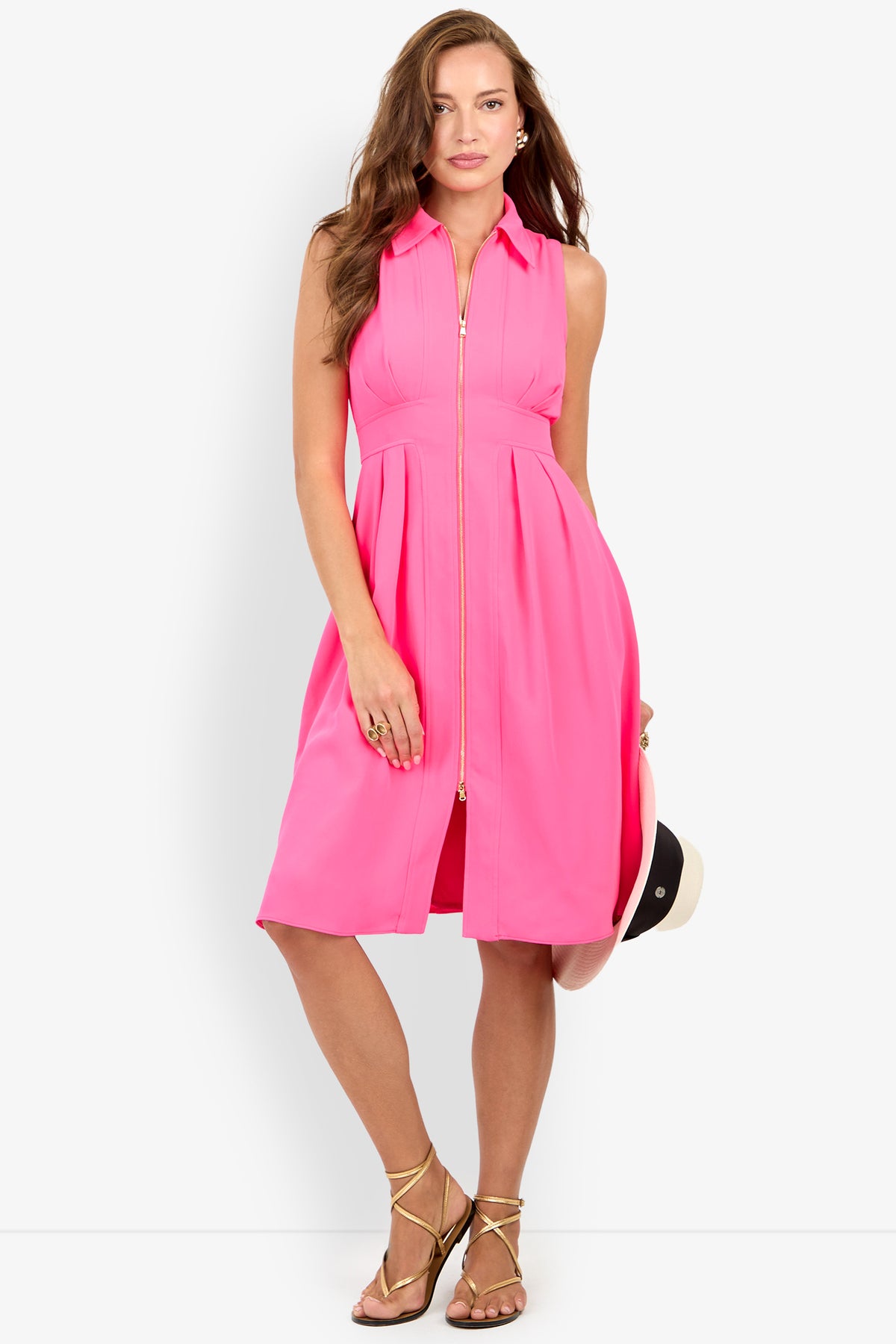 Woman in the Farfalla Sleeveless Midi Dress, a bright pink, knee-length dress with a front zipper, holding a hat and wearing gold sandals against a plain white background.