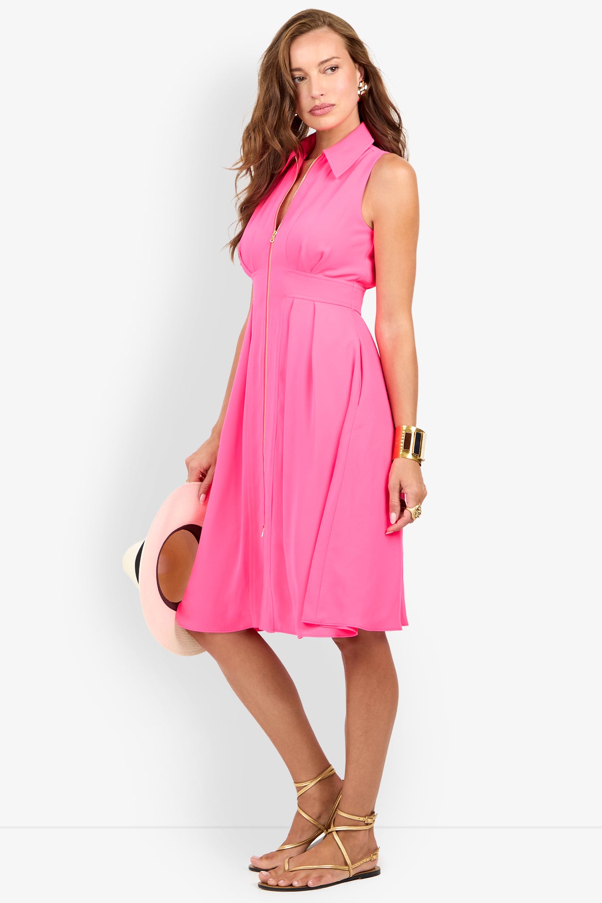 Woman in a Farfalla Sleeveless Midi Dress holds a hat by her side. She pairs the bright pink, collared dress with gold sandals, chunky bracelets, and rings against a plain white background.