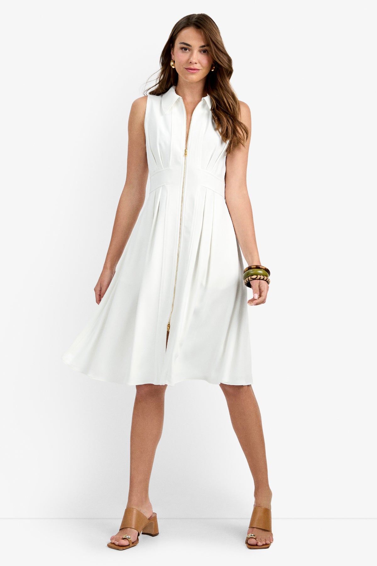 A woman models the Farfalla Sleeveless Midi Dress—collared, white, midi-length with a front zipper—paired with tan heeled sandals and bracelets on her right wrist, standing against a plain white background.