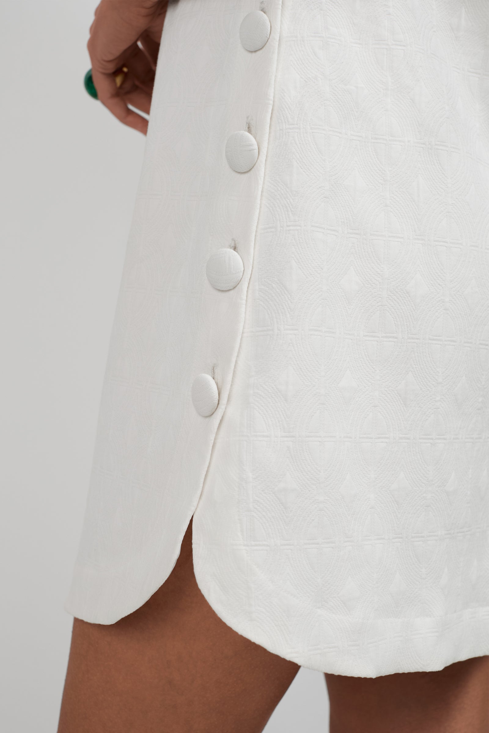 Close-up of the Atina Button Side Mini Dress, featuring white textured fabric, large fabric-covered side buttons, and a small hem slit, worn by a person with medium brown skin.