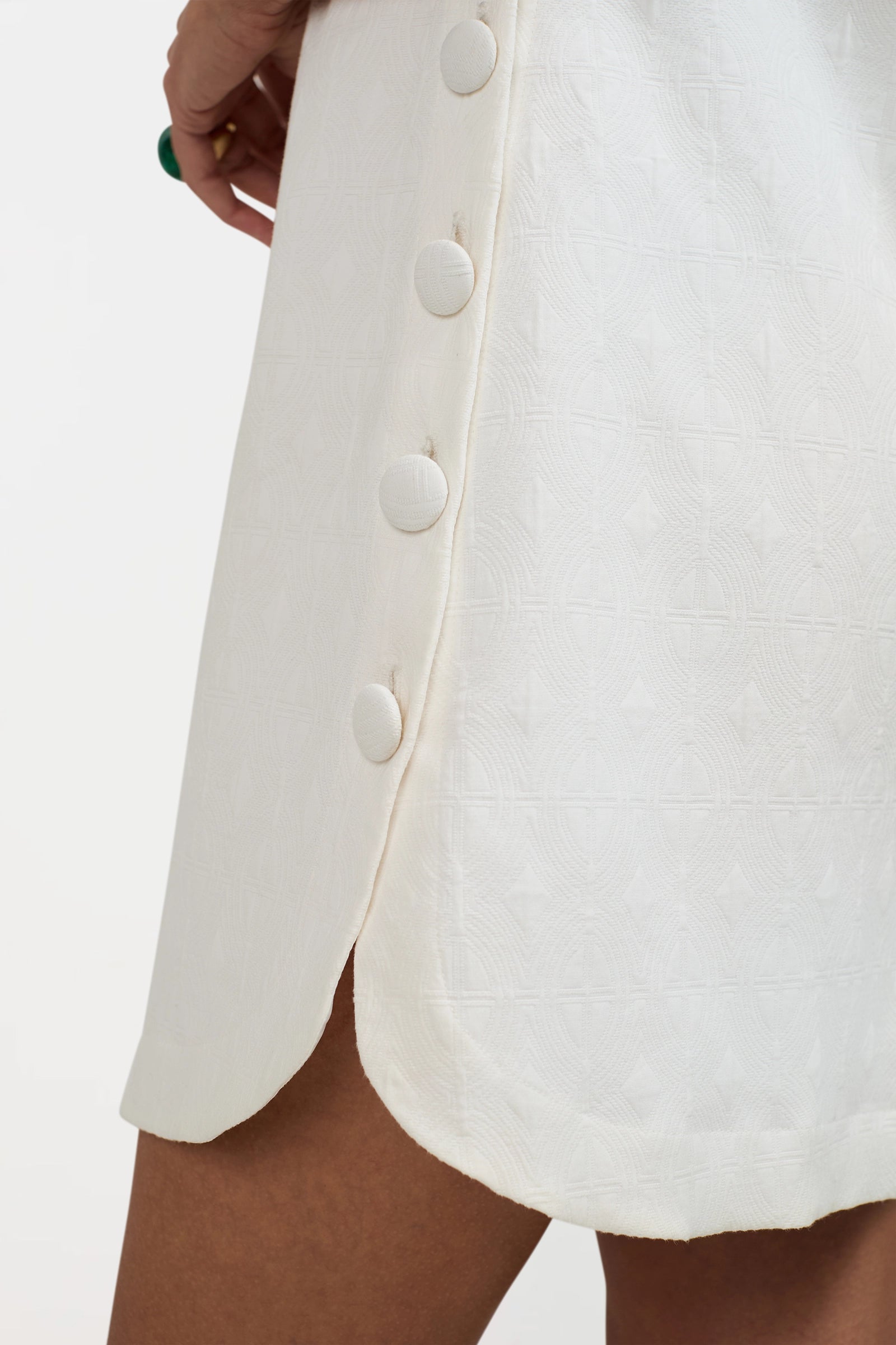 A close-up of the Atina Button Side Mini Dress in white, featuring large fabric-covered side buttons and a small hem slit, modeled on a person with medium skin tone.