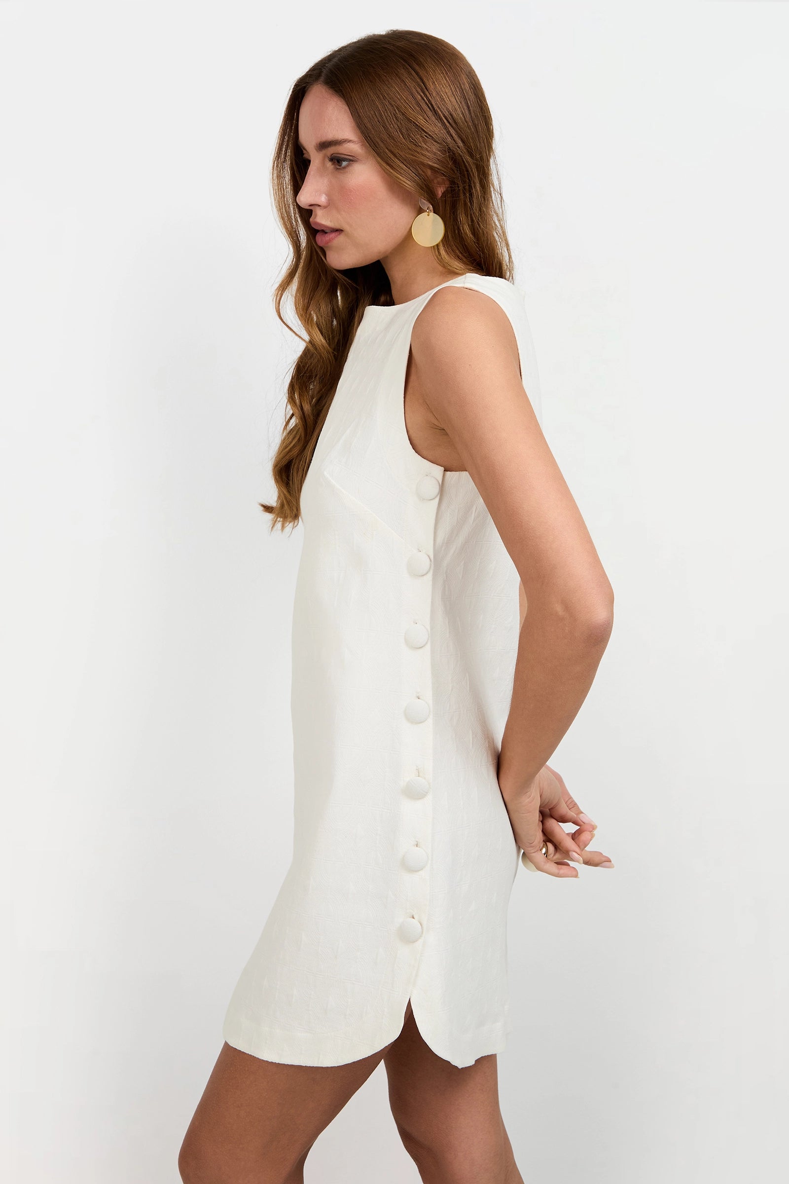 A woman with long, wavy brown hair models the Atina Button Side Mini Dress, a sleeveless white mini dress featuring large side buttons. She stands sideways against a plain white background, hands behind her back and looking ahead.