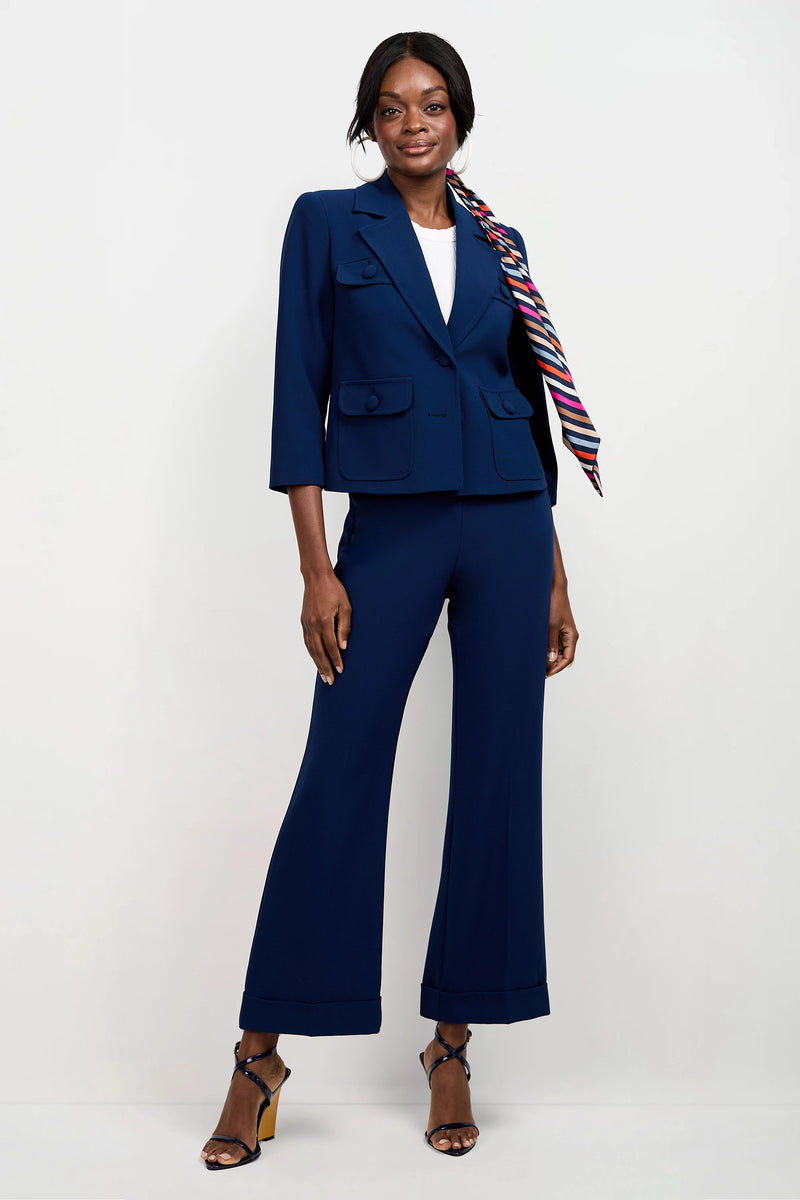 A woman stands against a white background wearing the Bicocca Patch Pocket Jacket in navy blue, paired with matching pants, a white top, strappy black heels, and a colorful striped scarf. She smiles confidently with one hand on her hip.