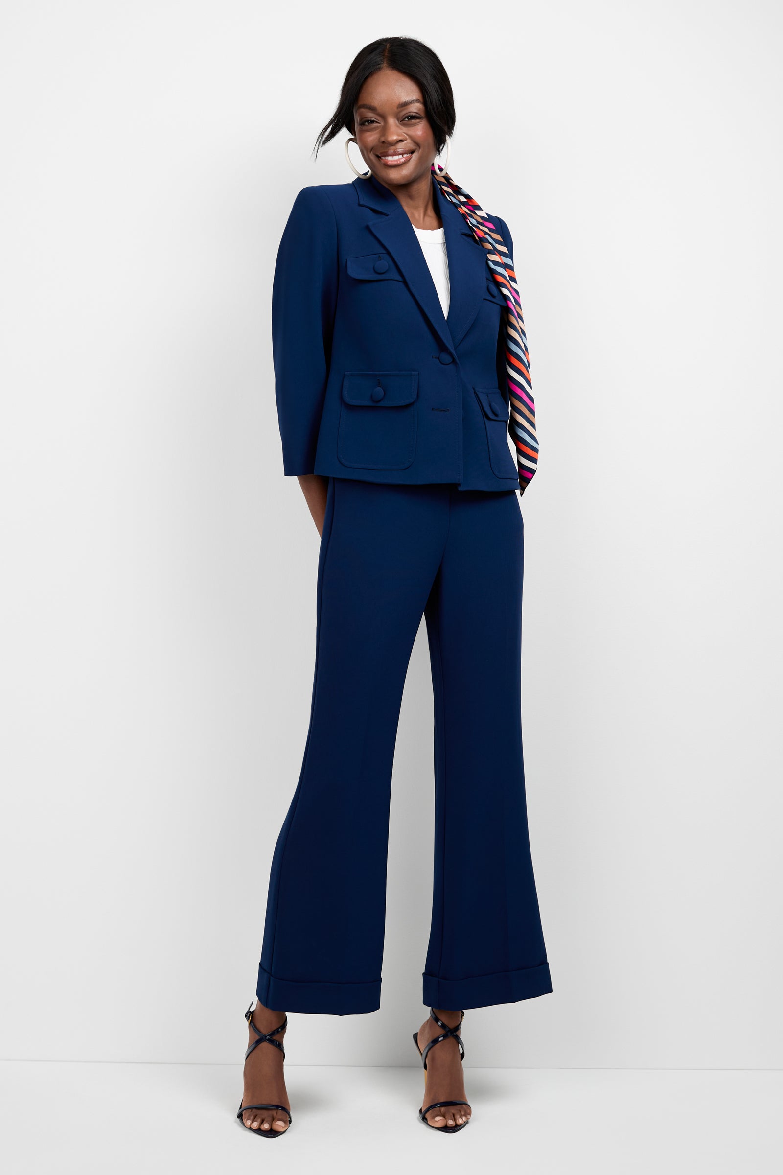 A woman smiles confidently against a white background, wearing the Bicocca Patch Pocket Jacket as part of a tailored navy pantsuit, paired with a white top, strappy black heels, and a colorful striped scarf in her ponytail.