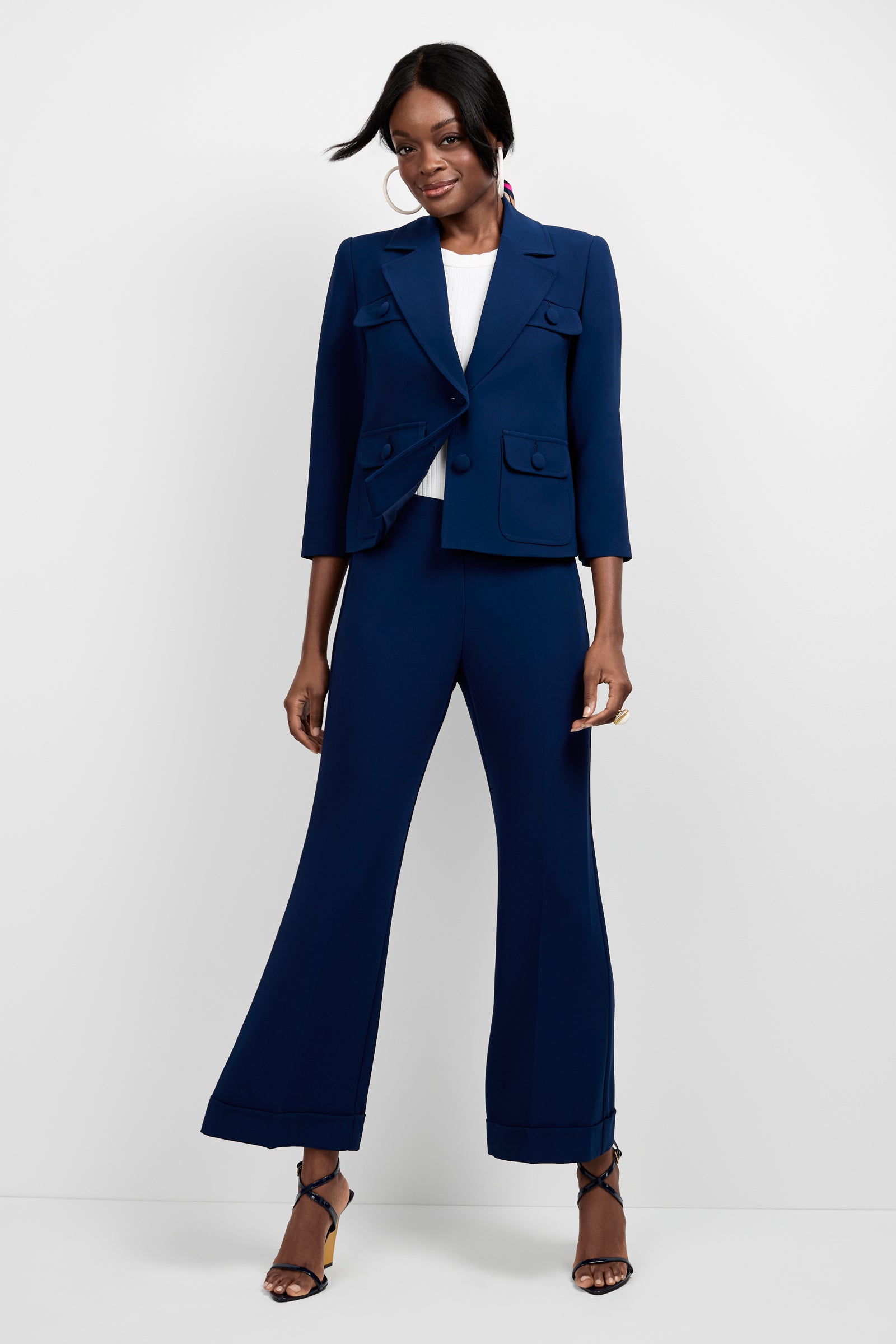 A woman stands against a white background wearing the Bicocca Patch Pocket Jacket—a tailored navy blue blazer—paired with wide-leg pants, a white top, and black heeled sandals. She smiles, hands relaxed by her sides.