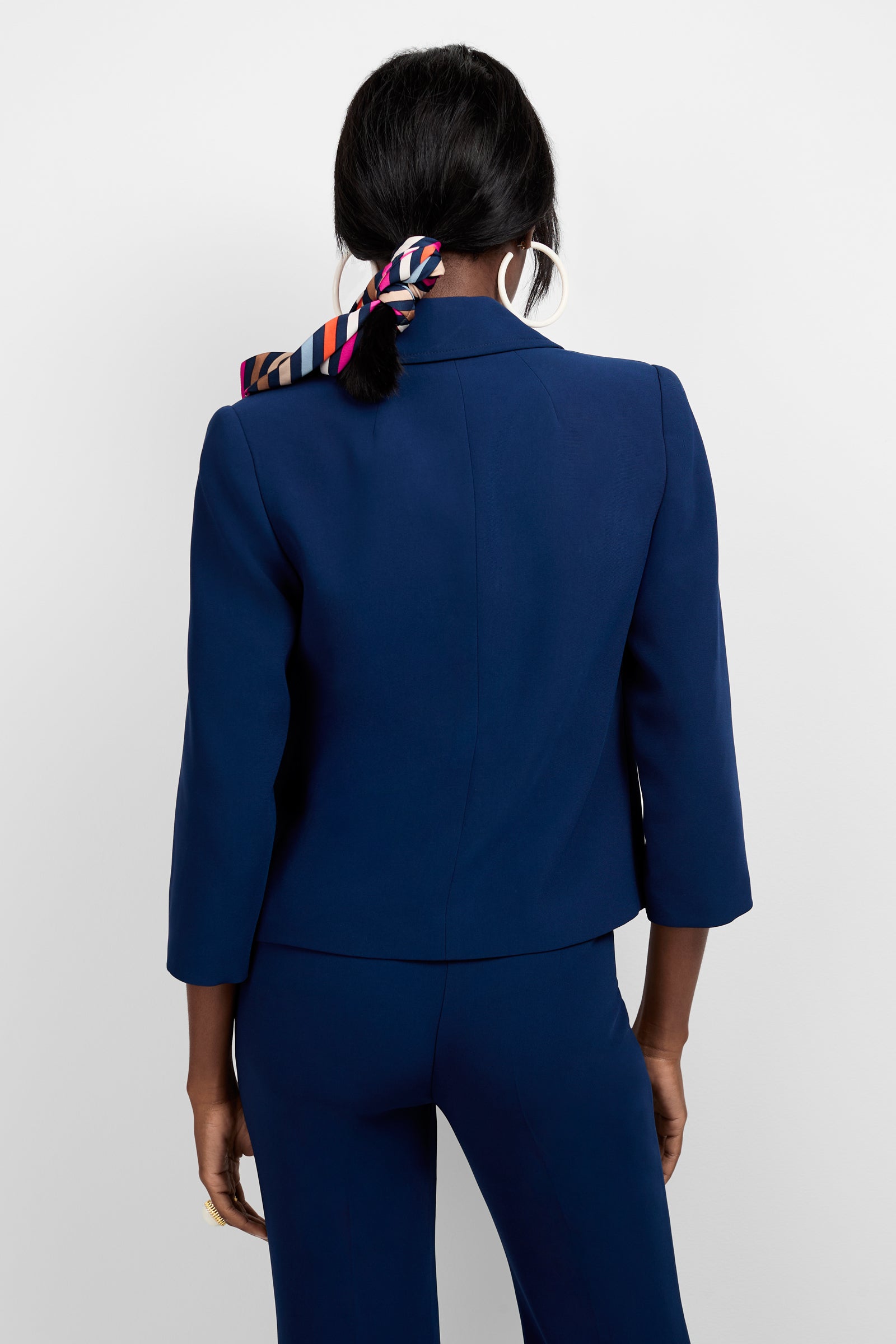 A woman faces away in the Bicocca Patch Pocket Jacket and matching blue pants. Her hair is tied with a striped scarf, and she wears large hoop earrings. The background is plain white.