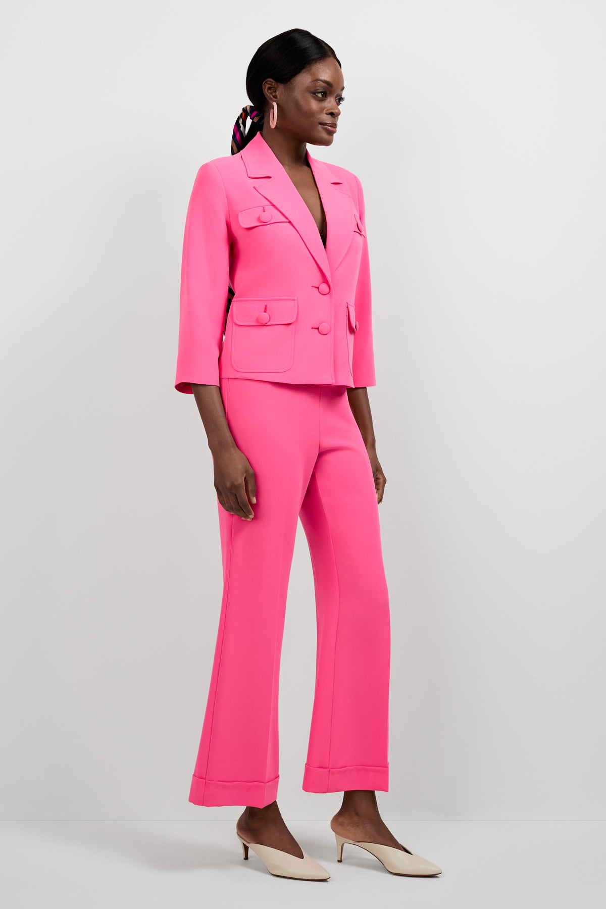 A woman stands against a plain background wearing the Bicocca Patch Pocket Jacket in bright pink with matching flared pants and white low-heeled shoes. Her hair is tied back with a scarf, and she looks slightly to the side.