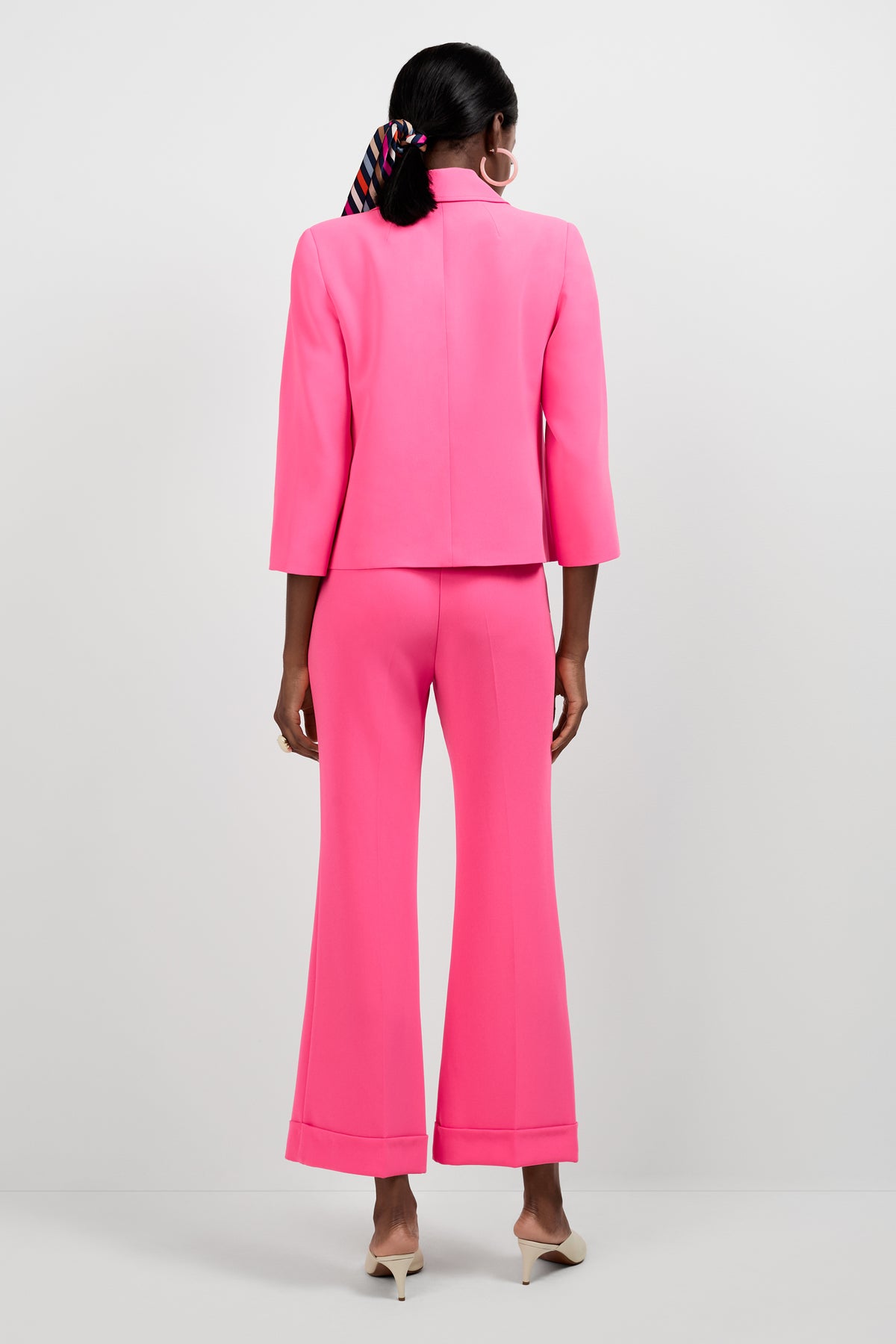 A woman faces away in the Bicocca Patch Pocket Jacket, a bright pink blazer with flared pants. She sports a striped hair scarf and white pointed heels against a plain white backdrop.
