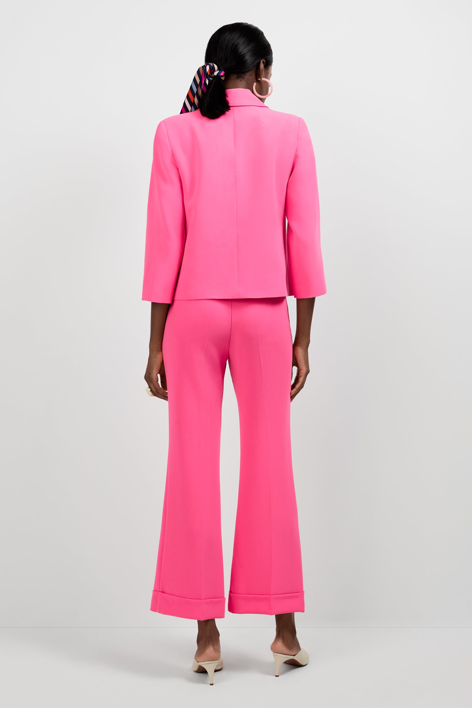 A woman faces away in the Bicocca Patch Pocket Jacket, a bright pink blazer with flared pants. She sports a striped hair scarf and white pointed heels against a plain white backdrop.