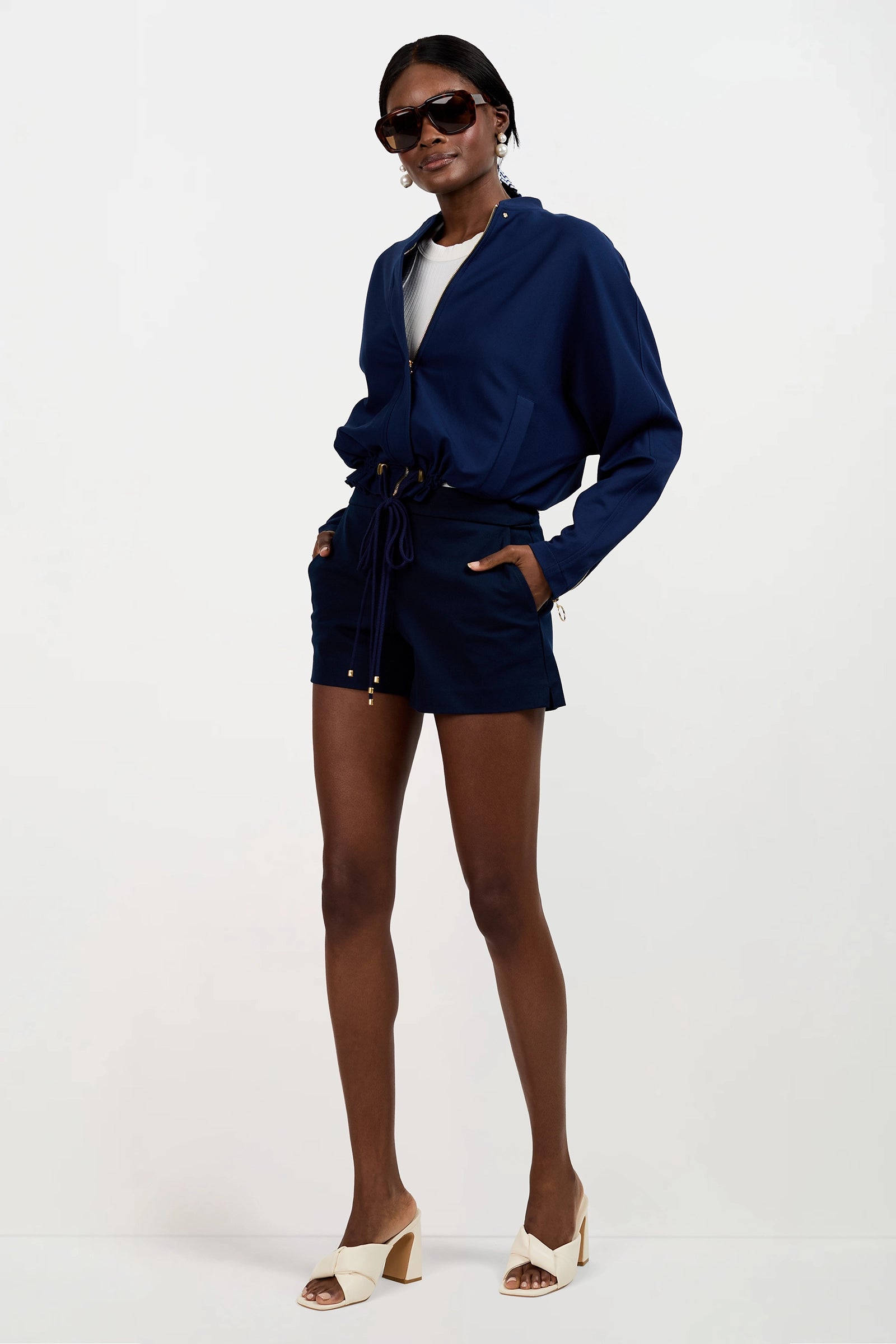 A woman stands with her hands in her pockets, wearing the Vignetta Cropped Jacket in navy blue with matching shorts, a white top, sunglasses, and beige heeled sandals against a plain white background.