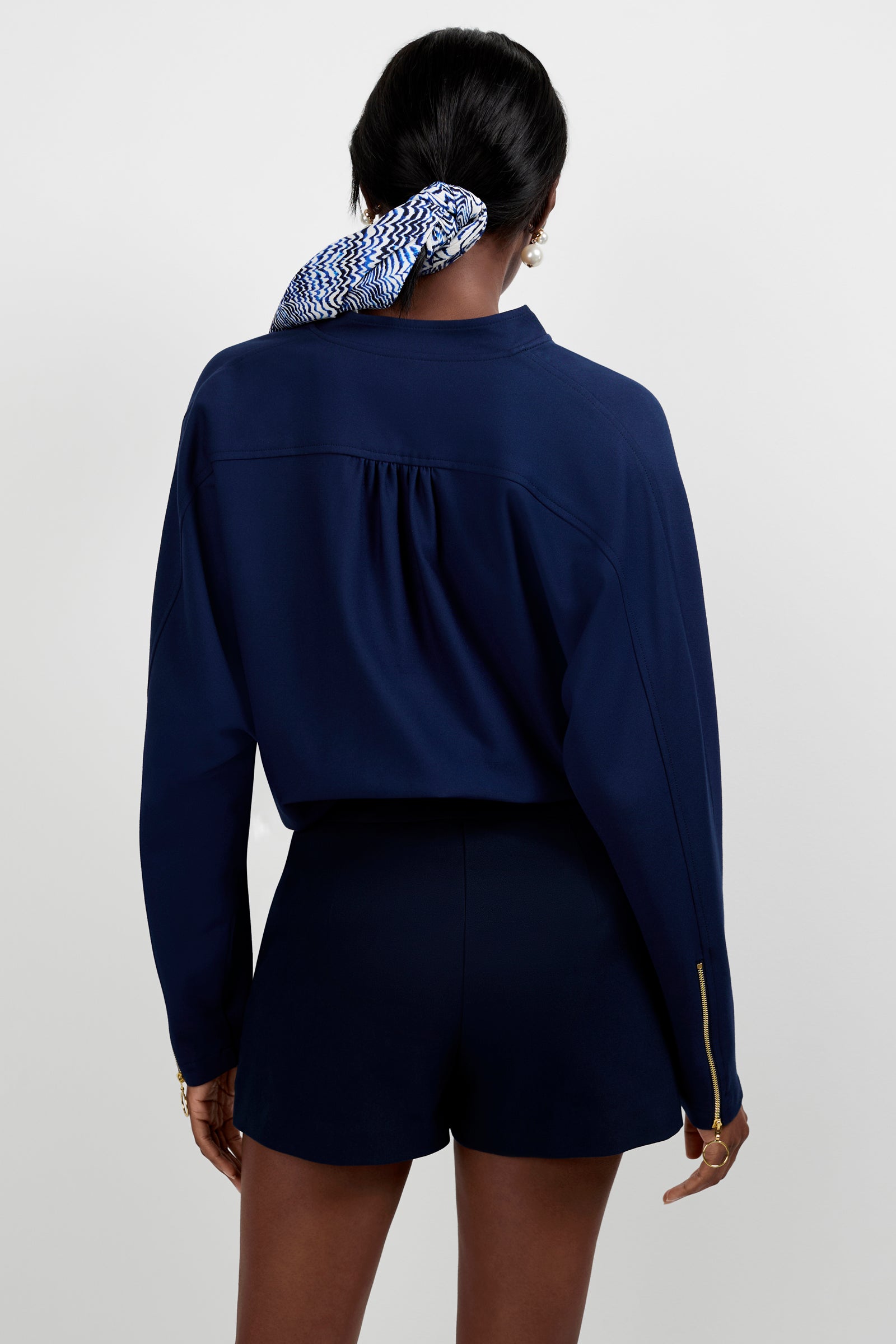 A woman with dark skin faces away, wearing the Vignetta Cropped Jacket in navy and matching shorts. Her hair is tied back with a blue and white patterned scarf against a plain, light-colored background.