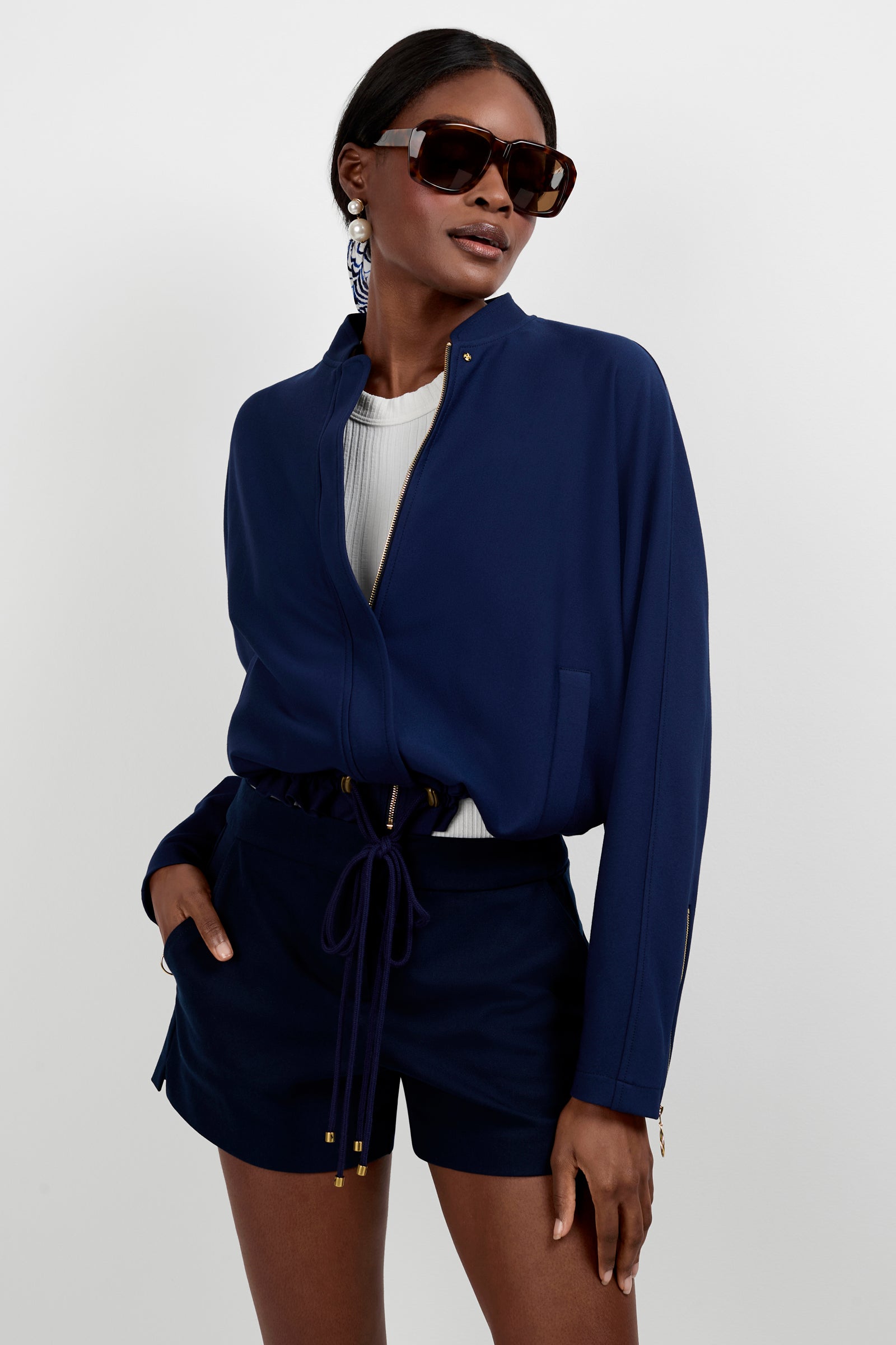 A woman poses confidently against a white background in the Vignetta Cropped Jacket in INK, paired with matching shorts, a white top, dark sunglasses, and large earrings, with one hand in her pocket.