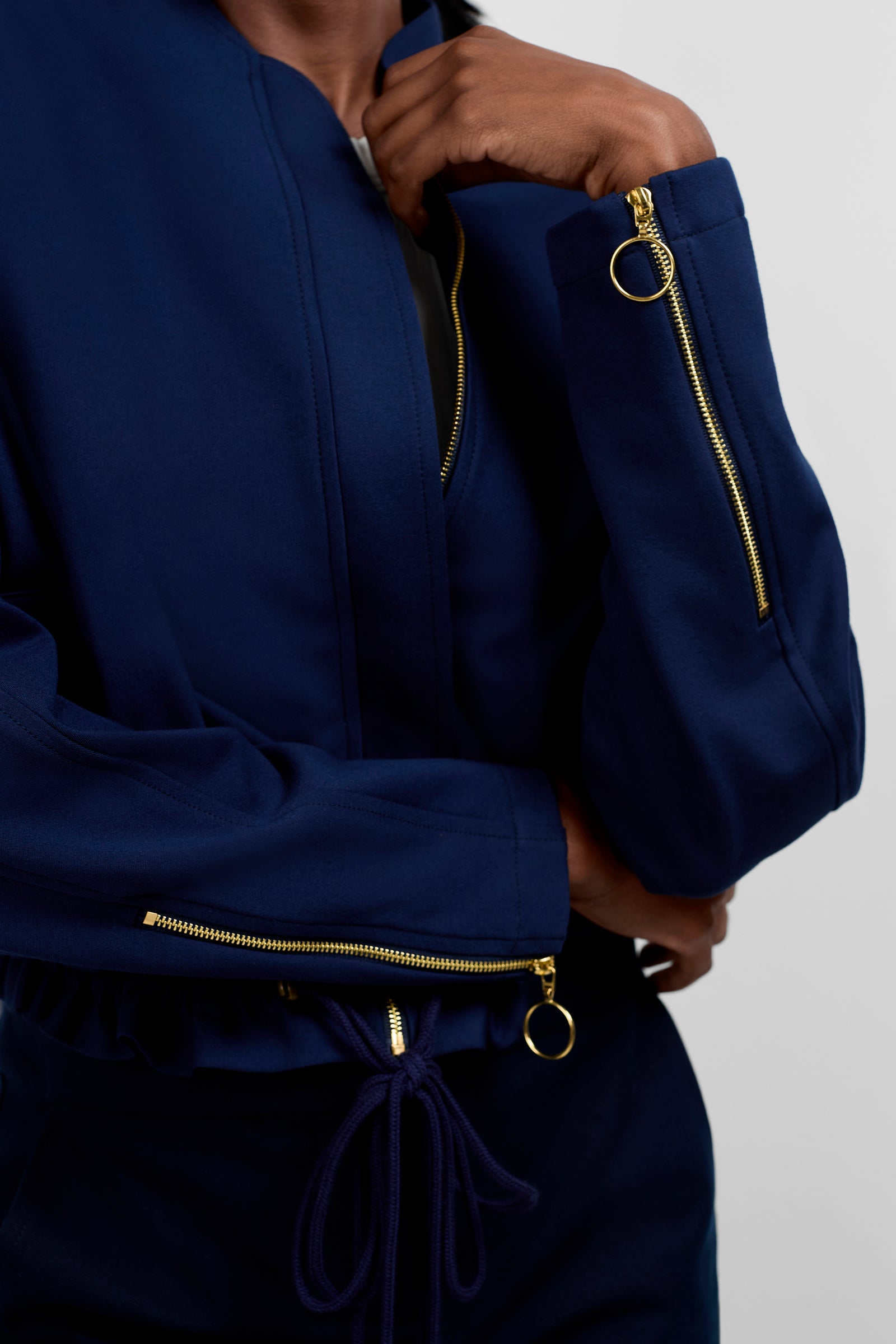 A person wears the Vignetta Cropped Jacket in navy with gold zipper details on the sleeves and front, paired with navy drawstring-waist pants. Their face is partially out of frame.