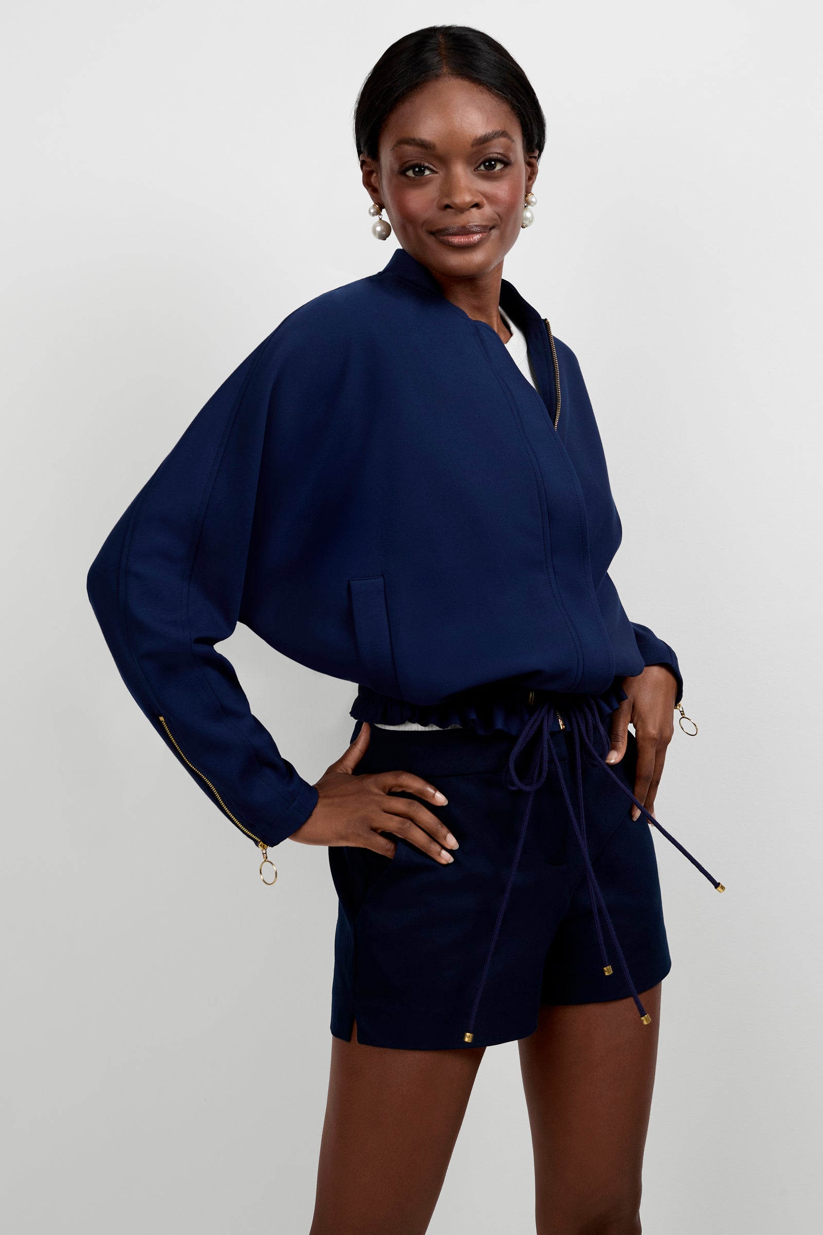 A woman stands confidently against a plain background wearing the Vignetta Cropped Jacket in navy blue with gold zipper details, paired with matching navy blue shorts. She poses with one hand on her hip and a slight smile.