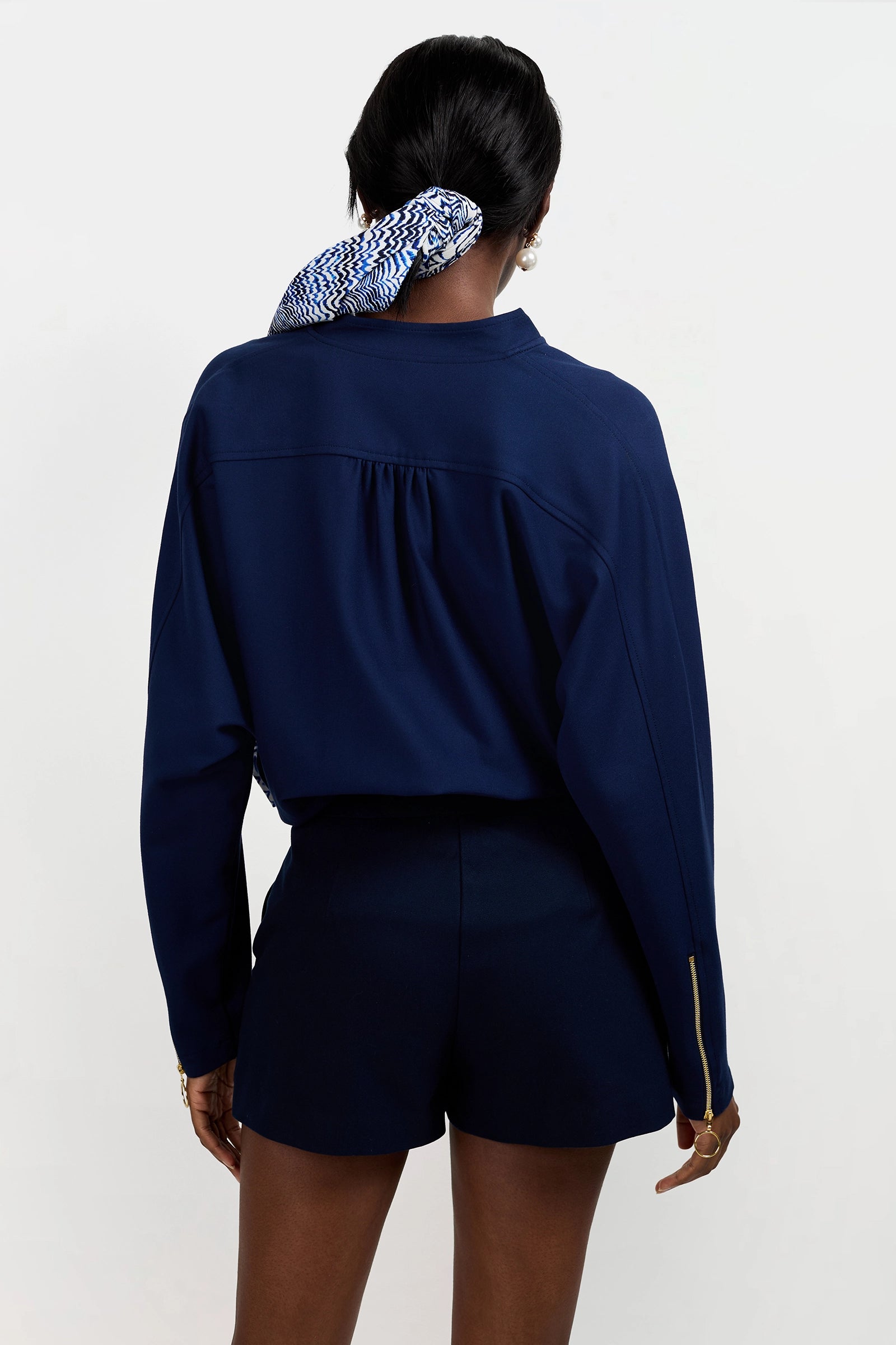 A woman stands facing away, wearing the Vignetta Cropped Jacket in navy with gold zipper details and matching shorts. Her hair is tied back with a white and blue patterned scarf, set against a plain white background.