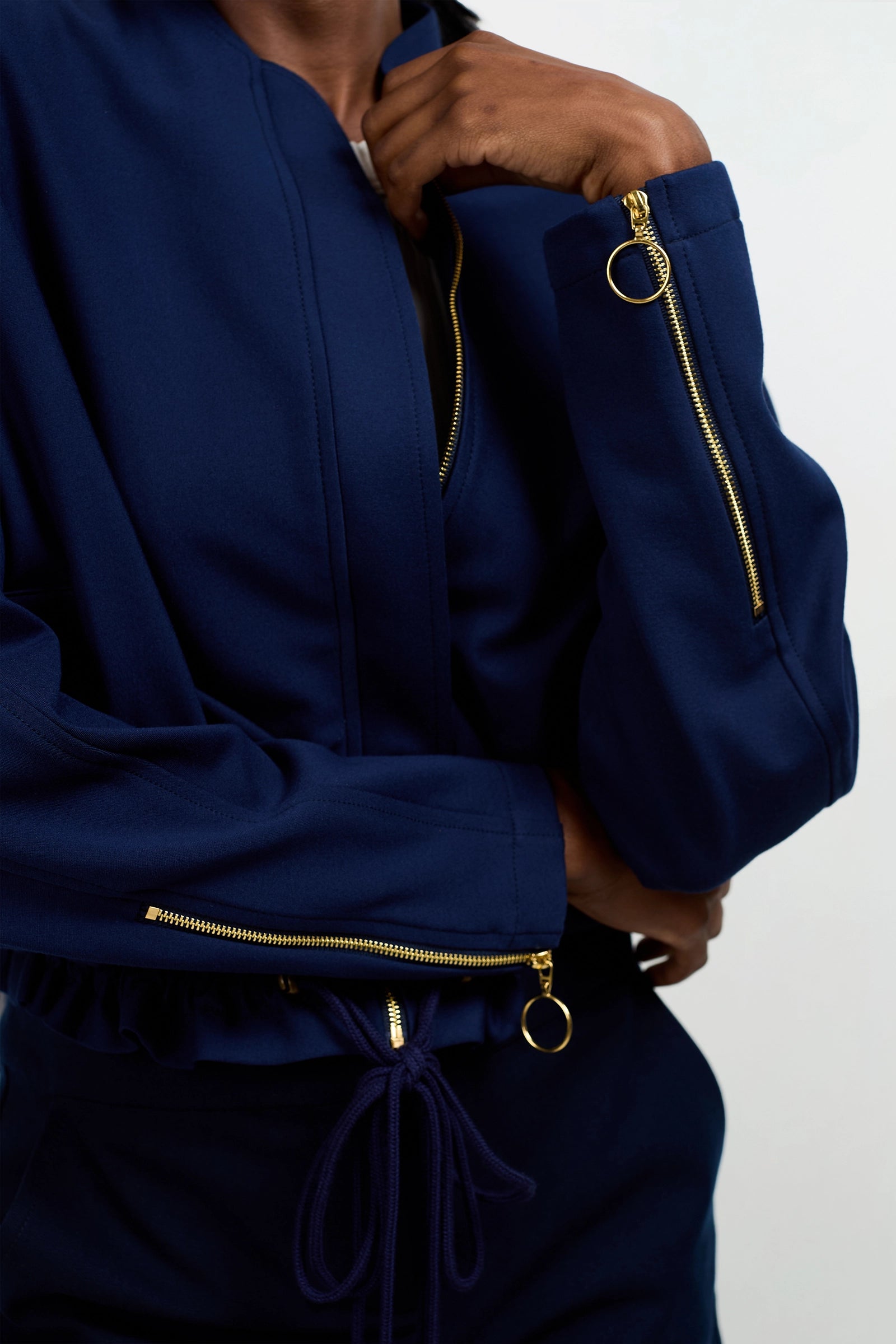A person wearing the Vignetta Cropped Jacket in navy blue with gold zippers and ring pulls, standing with arms crossed. The focus is on the sleeve and zipper details.