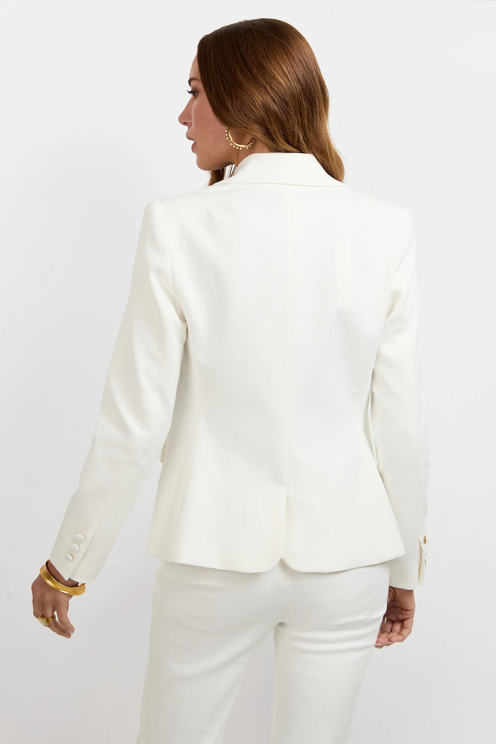 A woman with long brown hair wears the Incanta Fitted Blazer and matching pants, standing with her back to the camera against a plain white background. She accessorizes with gold hoop earrings and a gold bracelet.