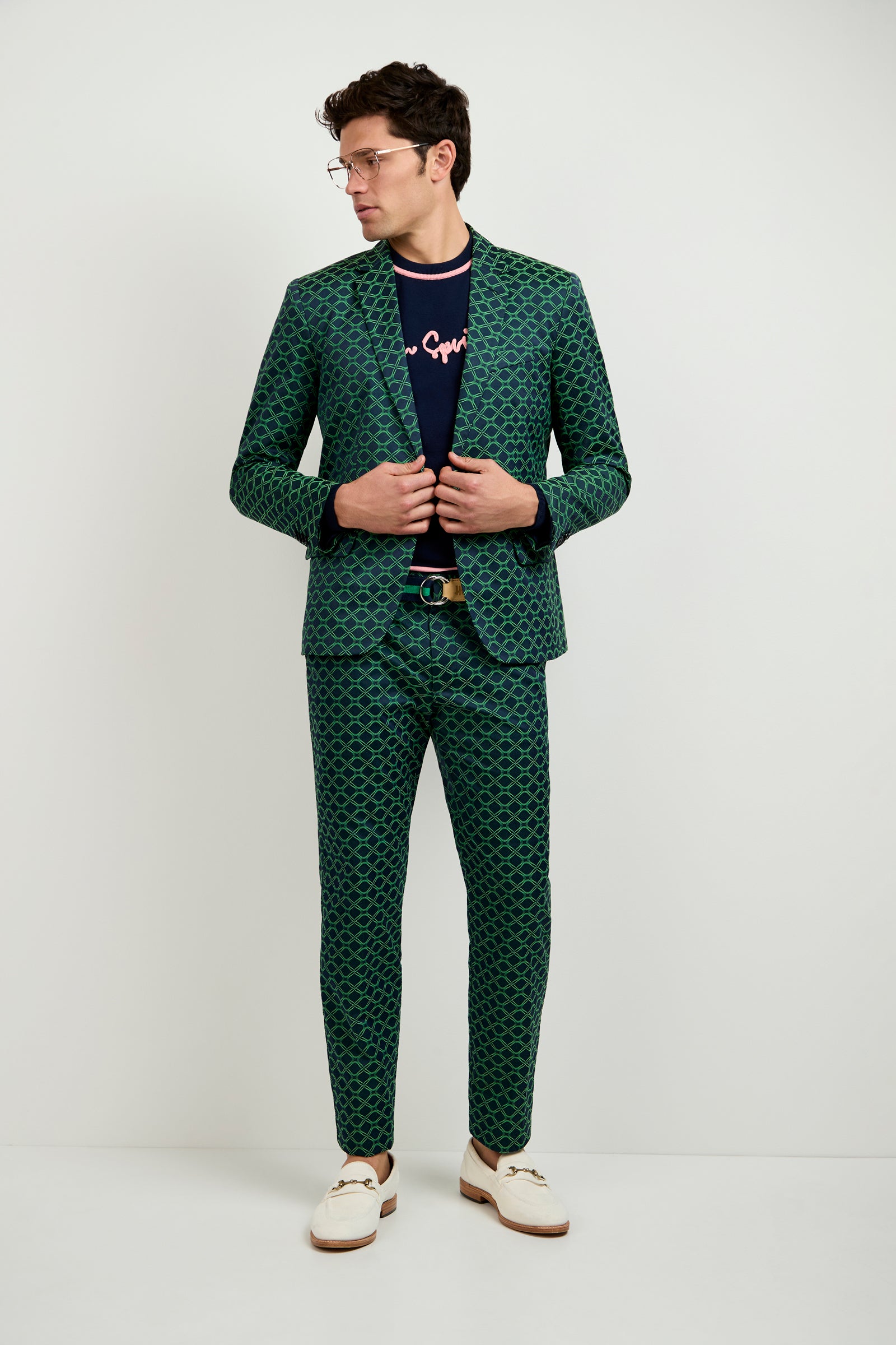 A man stands against a white wall wearing the Thurston Blazer in Clover Geo Jacquard, a navy text shirt, brown belt, white loafers, and glasses. He looks to the side while adjusting his jacket.