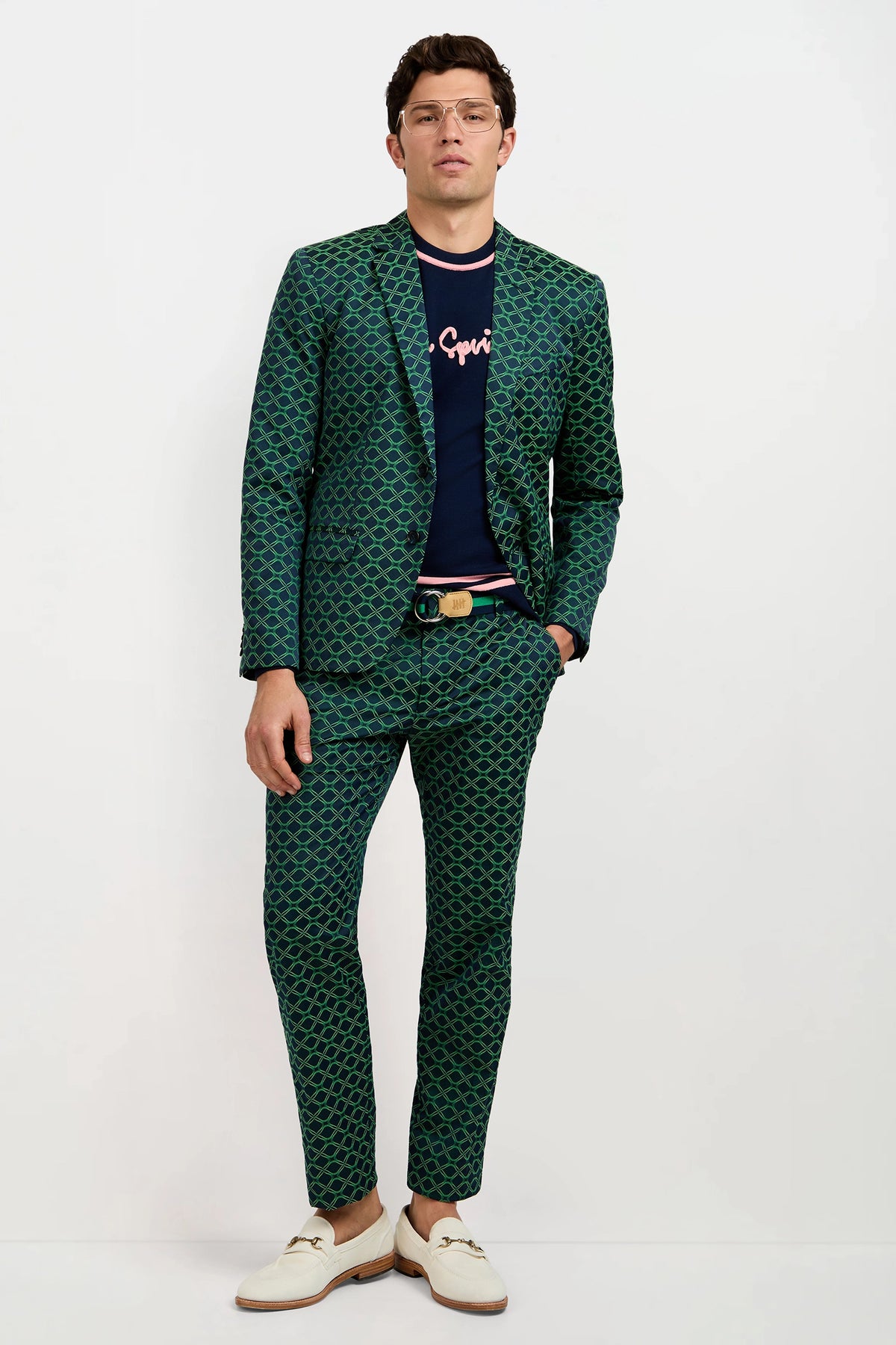A man wearing a green patterned suit, the Palm Springs Crewneck Sweater, white loafers, glasses, and a tan belt stands against a plain white background.