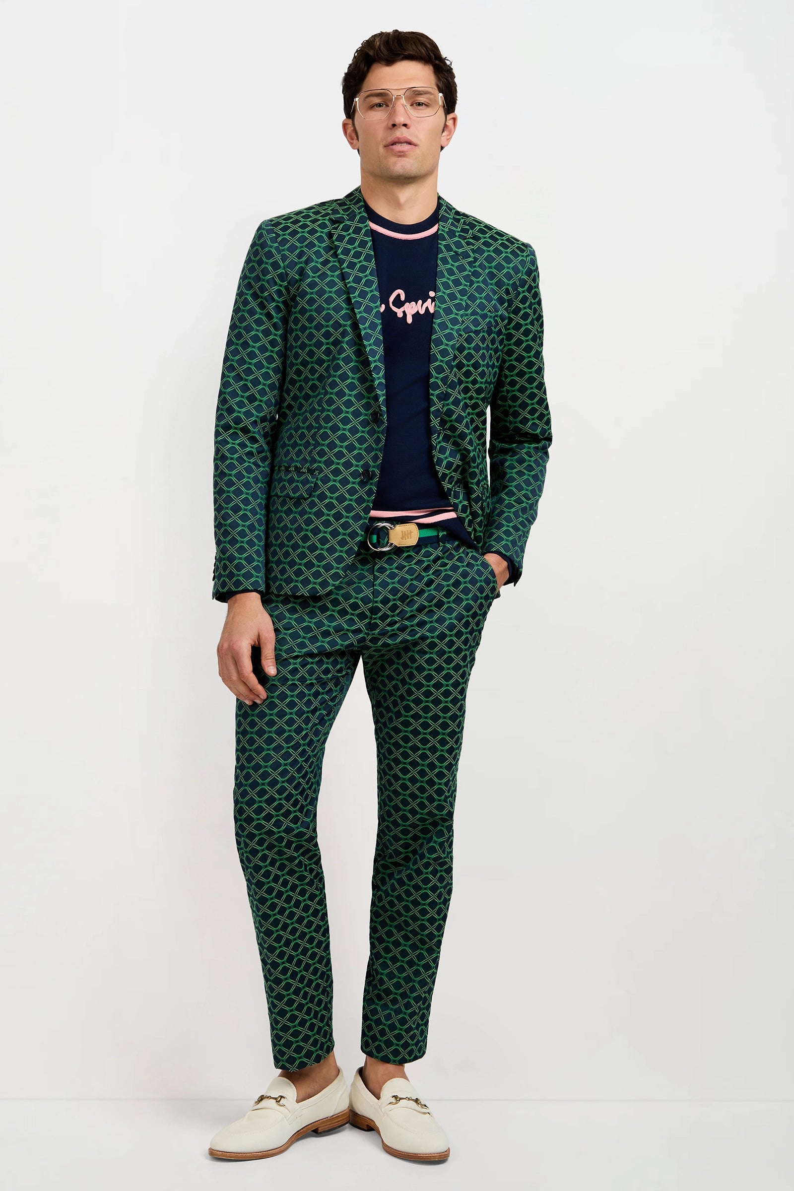 A man wearing a green patterned suit, the Palm Springs Crewneck Sweater, white loafers, glasses, and a tan belt stands against a plain white background.