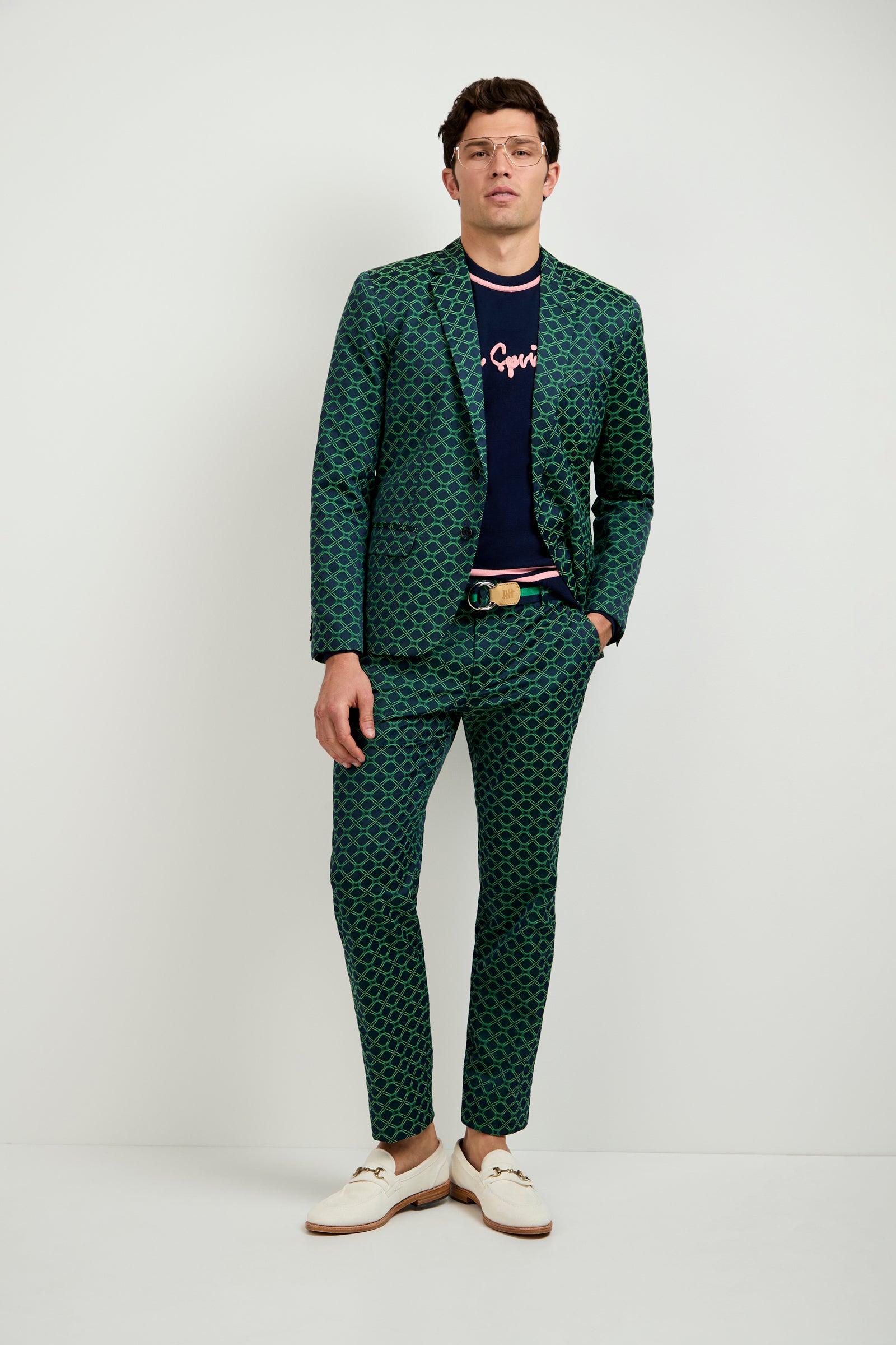 A man stands against a plain white background, wearing the Clyde Slim Trouser in Clover Geo Jacquard, a navy graphic t-shirt, cream loafers, glasses, and a belt, confidently looking at the camera with one hand in his pocket.