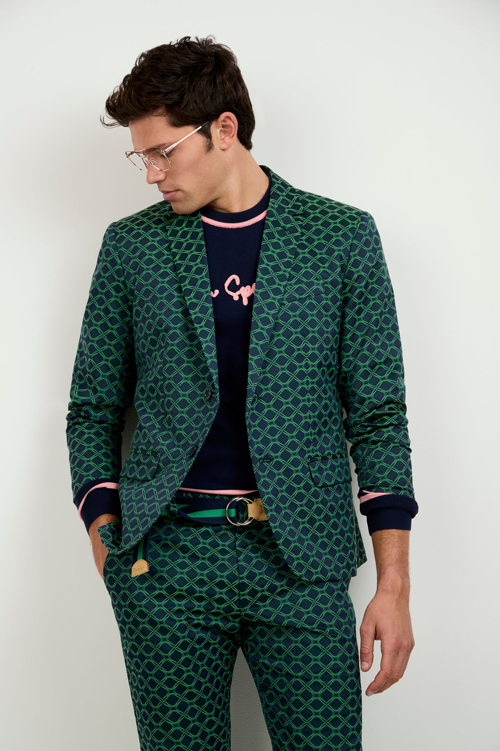 A man wearing glasses stands against a white background, looking down. He wears the Thurston Blazer - Clover Geo Jacquard with matching pants over a navy sweater featuring pink trim and lettering.