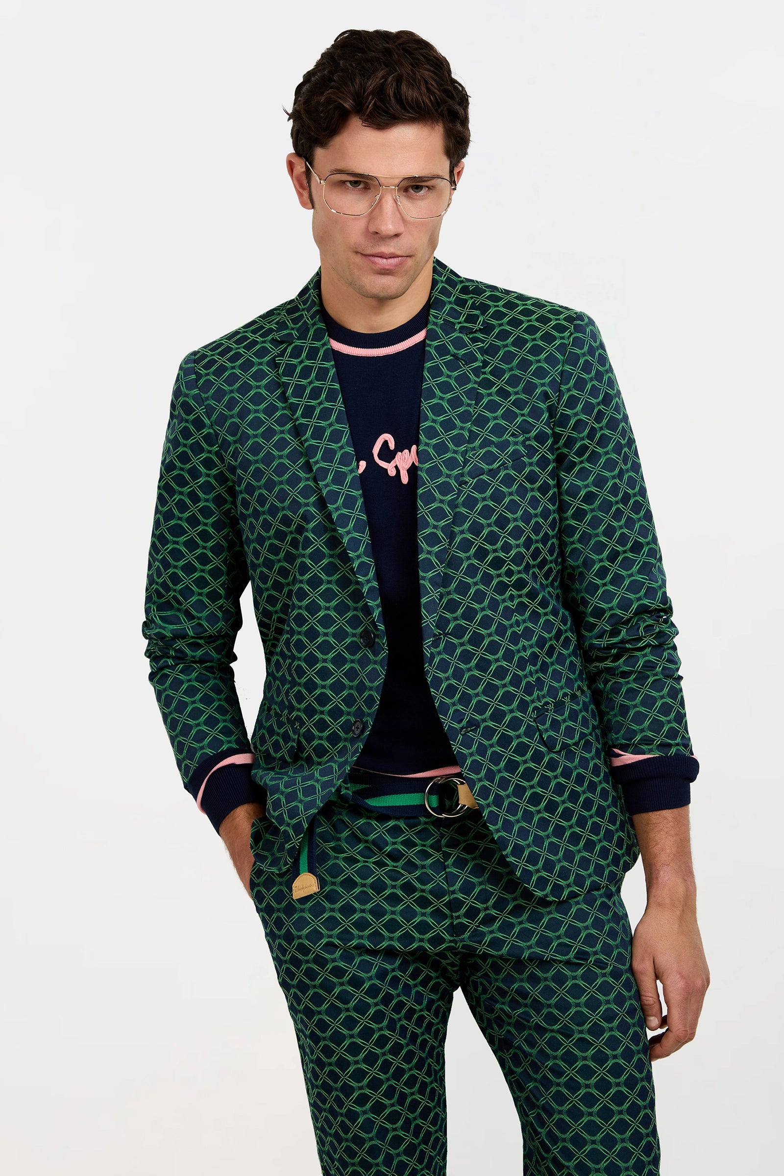 A man wears the Thurston Blazer in Clover Geo Jacquard, styled with a navy sweater featuring pink writing, a gold-buckle belt, and thin glasses, posing with one hand in his pocket against a plain background.