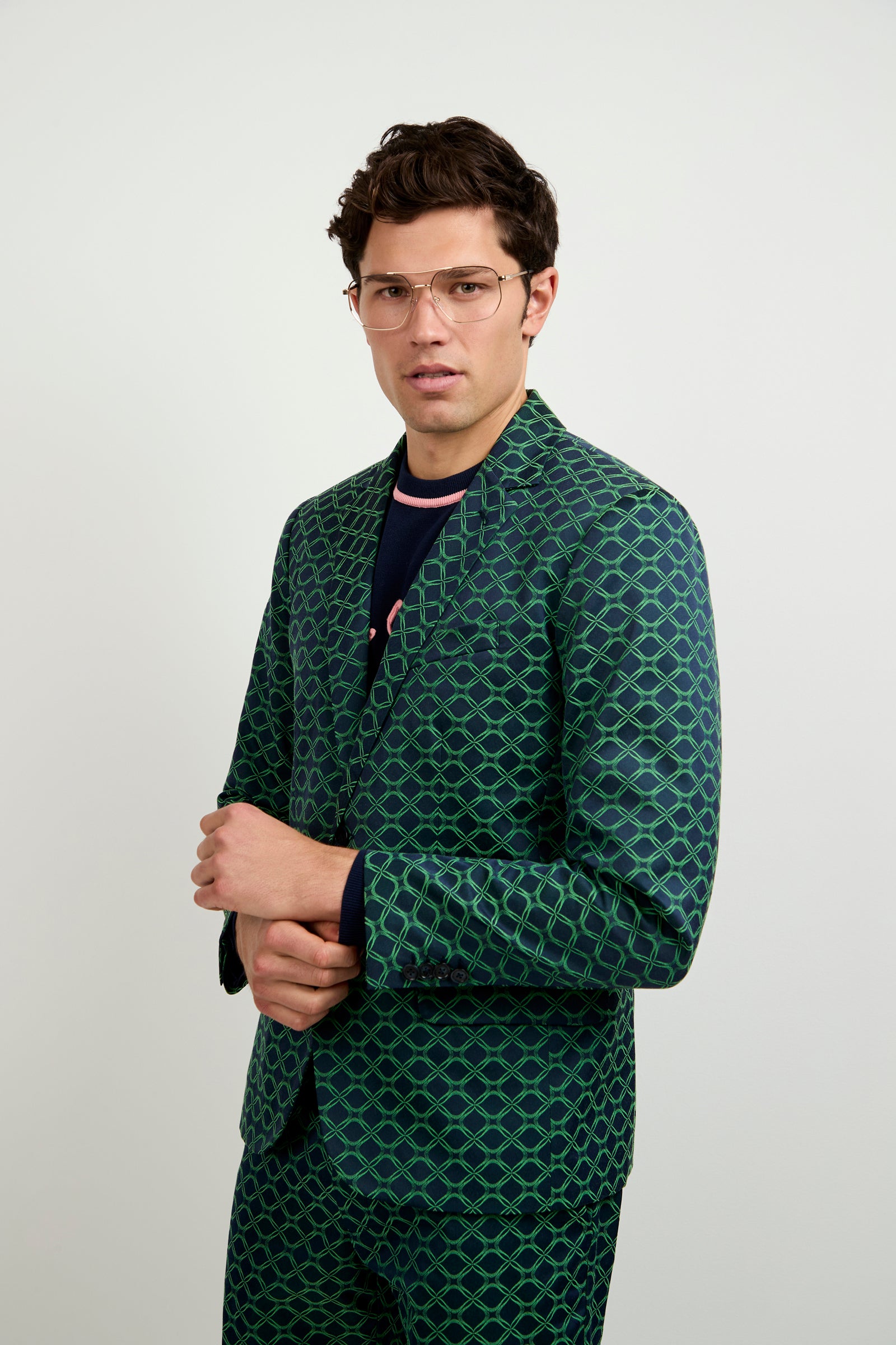 A man with short dark hair and glasses wears the Thurston Blazer in Clover Geo Jacquard, posing with one hand holding his opposite arm against a plain light background.