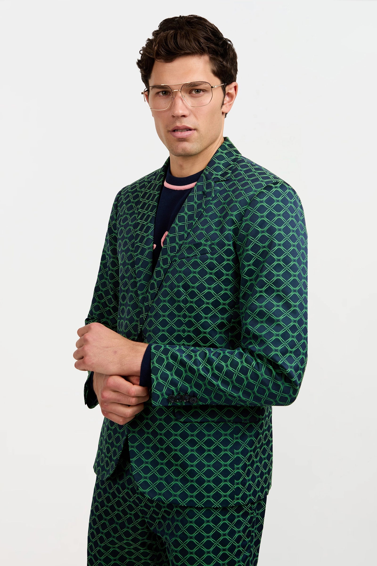 A man in the Thurston Blazer - Clover Geo Jacquard, featuring glasses and a green suit with a blue geometric pattern, stands confidently against a plain white background.