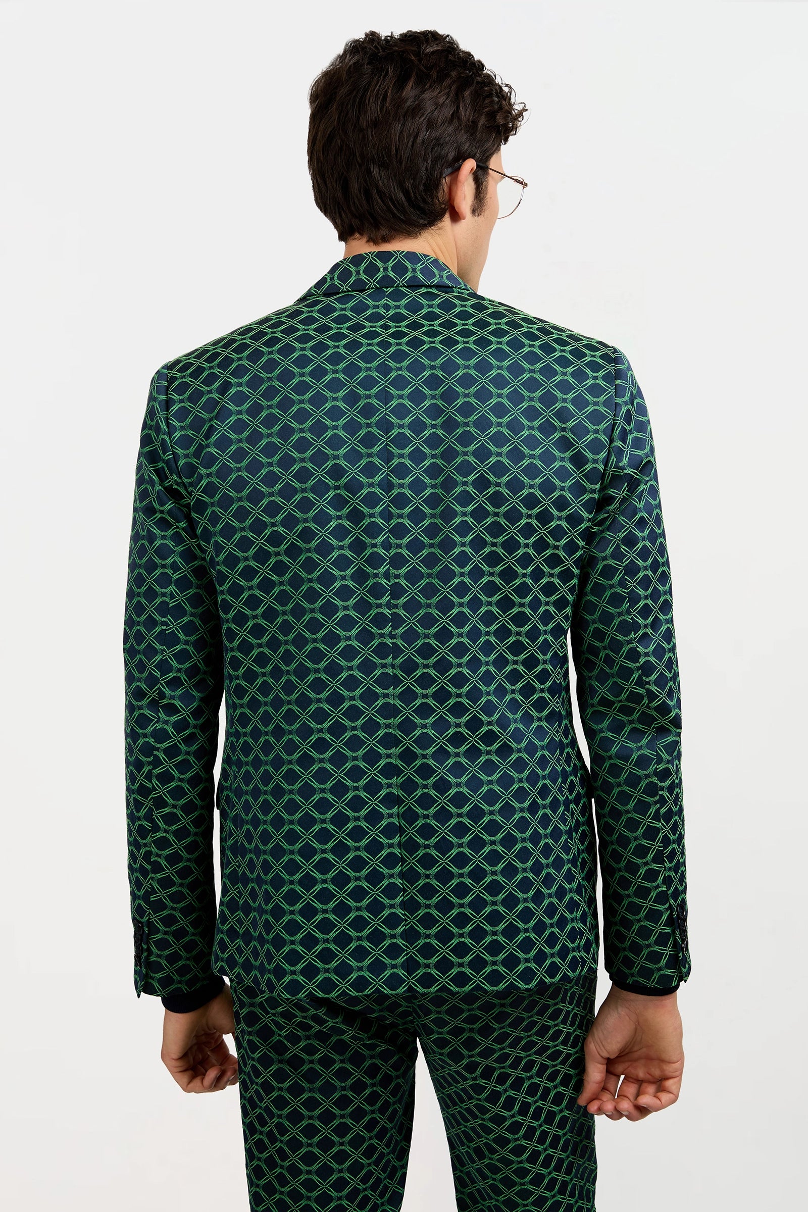 A man faces away from the camera wearing the Thurston Blazer in Clover Geo Jacquard, featuring a dark green base with a bright green geometric pattern, set against a plain white background.