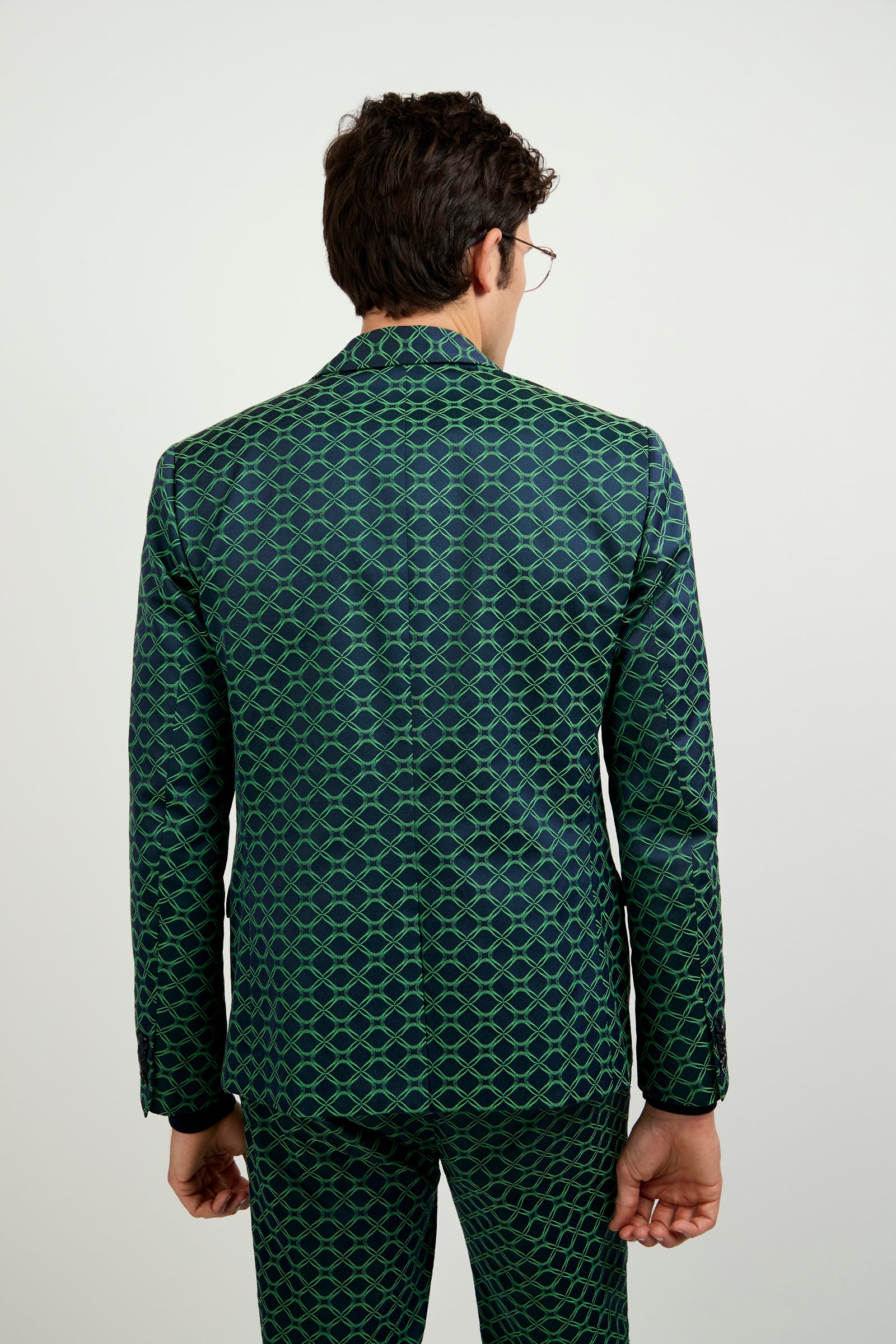 A person with short dark hair and glasses, dressed in the Thurston Blazer – Clover Geo Jacquard, stands with their back to the camera against a plain light background.