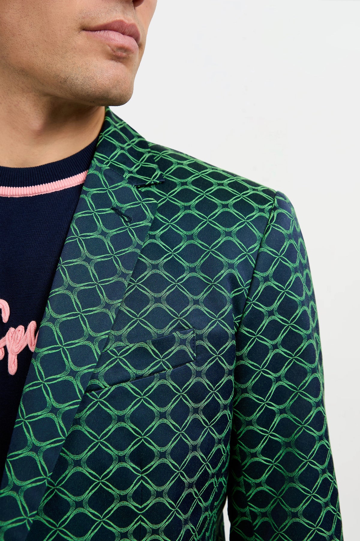 A man wears the Thurston Blazer in Clover Geo Jacquard—a dark blazer with a bold green geometric pattern—over a navy sweater with pink lettering, shown from the shoulders up against a plain white background.