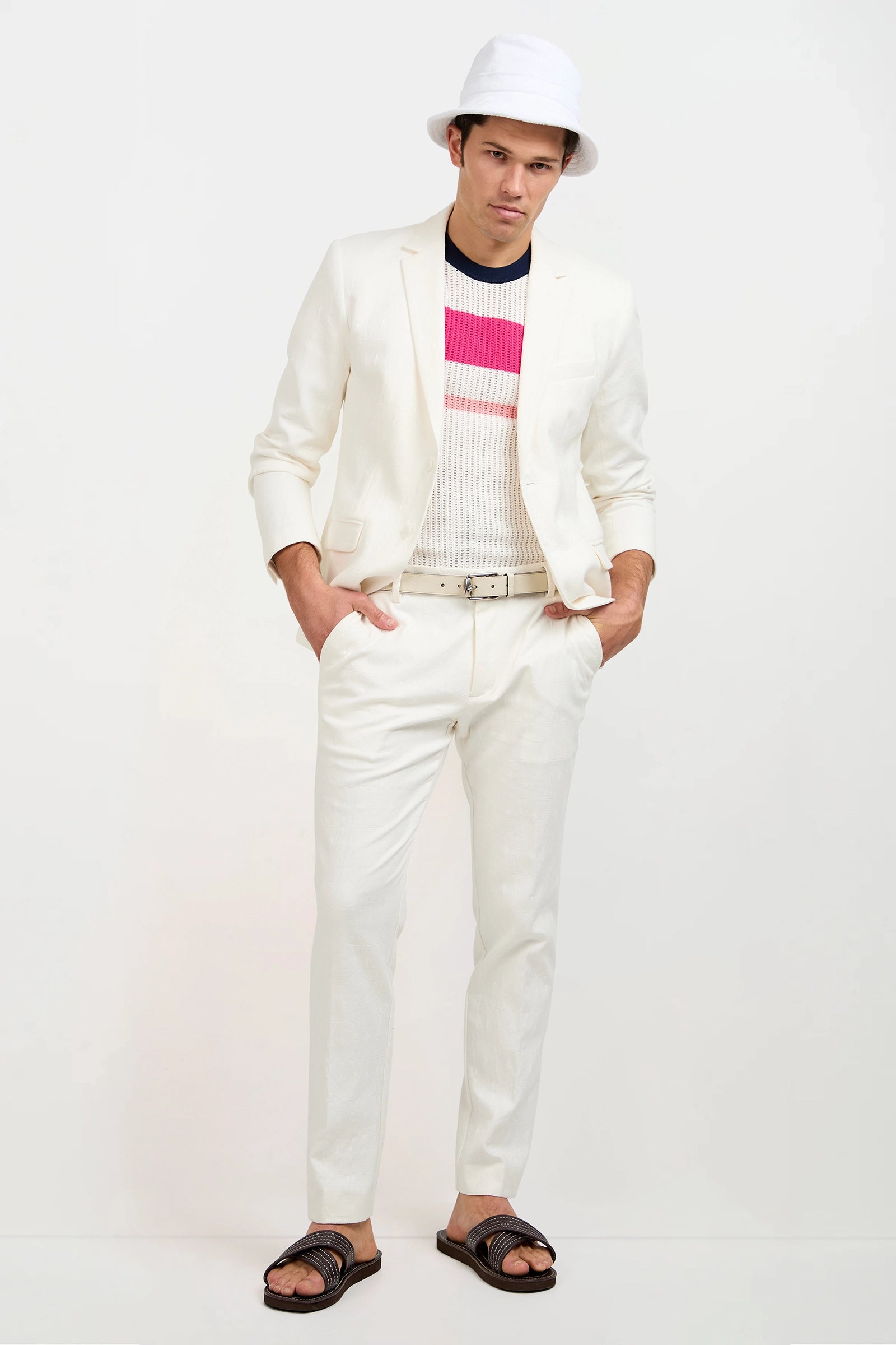 A man stands facing the camera, hands in pockets, wearing the Clyde Slim Trouser in Tegola Textured Jacquard with a white suit, striped sweater, white belt, bucket hat, and dark sandals against a plain background.