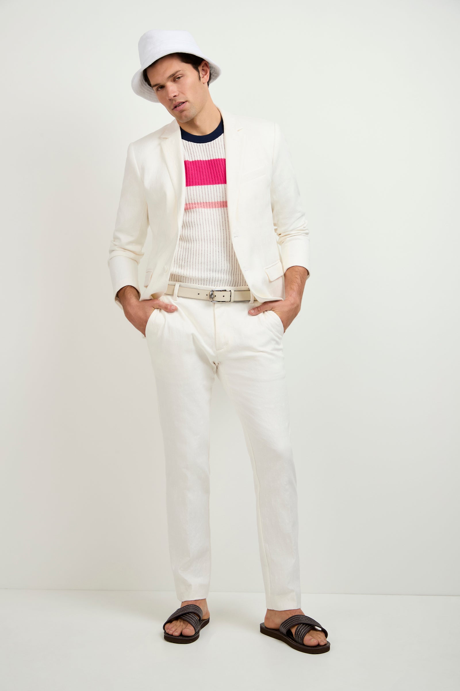 A man stands against a white background wearing Clyde Slim Trouser - Tegola Textured Jacquard, a white suit, bucket hat, striped knit shirt, white belt, and black sandals, with his hands in his pockets.